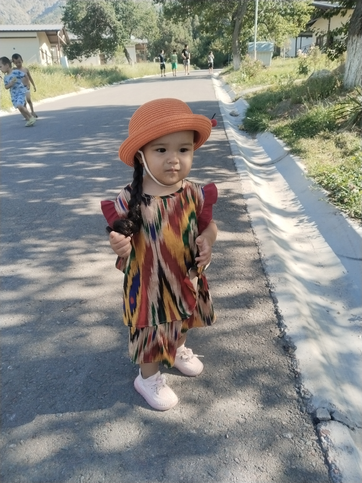 A young child wearing a colorful ikat print dress and an orange hat stands on a paved road. The child has a single braid extending from under the hat. In the background, other children are visible walking and playing on the road, and buildings and trees line the sides of the road. The scene appears to be in a residential area of Akata, Uzbekistan, possibly a park or neighborhood street. Sunlight casts shadows on the road.