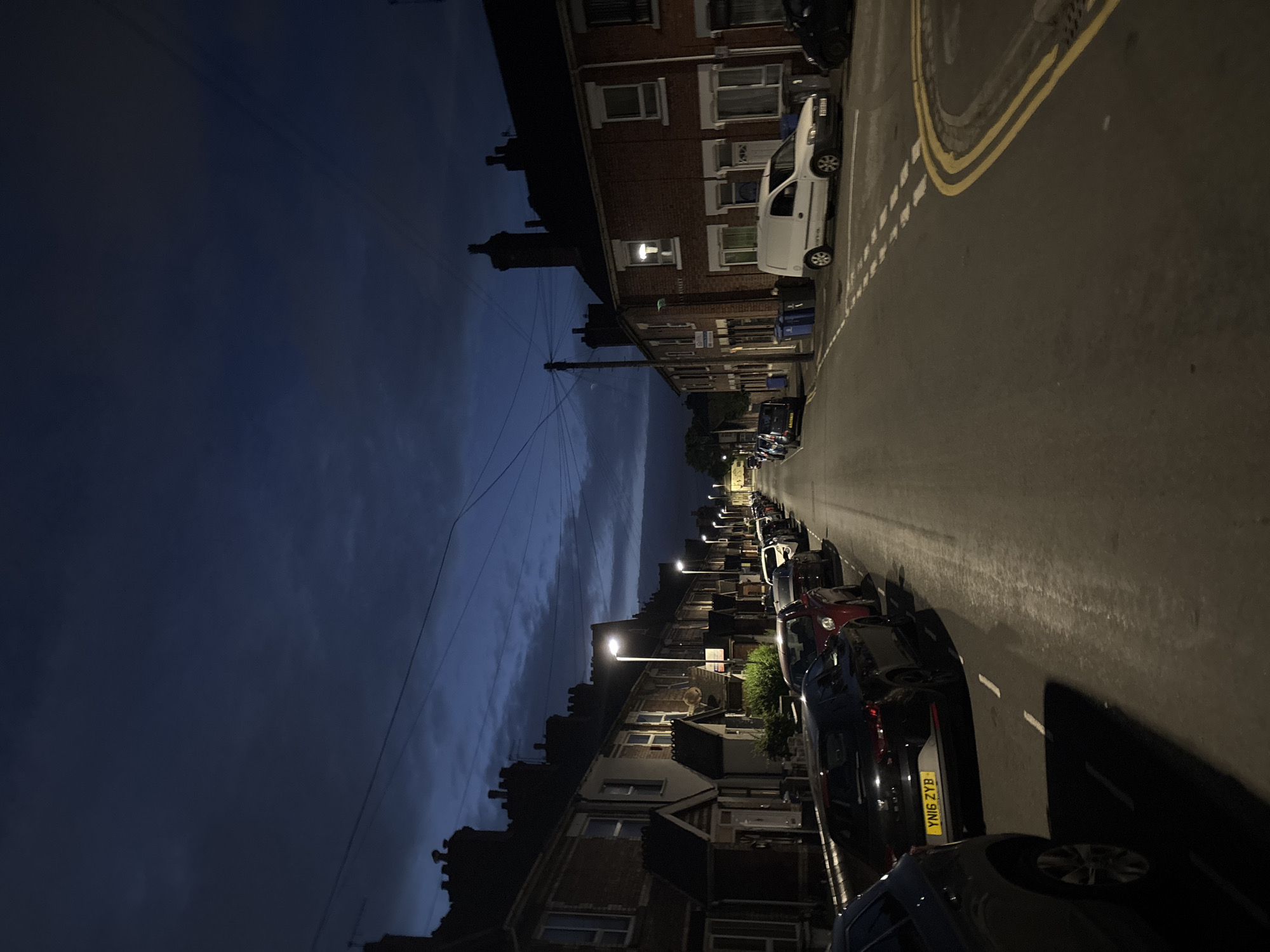 The image depicts a residential street in Stoke-on-Trent, United Kingdom, at dusk. The scene is dominated by terraced houses lining both sides of the street, with parked cars filling the available spaces along the road. Streetlights cast a warm glow on the asphalt, illuminating the parked vehicles and the pavement. The sky above is a deep blue, with scattered clouds suggesting the transition from day to night. The overall atmosphere is quiet and still, characteristic of a residential neighborhood in the evening. There are no people visible in the image. The houses appear to be made of brick, with typical pitched roofs and chimneys. Some windows are lit, suggesting that residents are at home. The street is paved and appears to be in good condition. The parked cars are a variety of makes and models, indicating a typical urban setting.