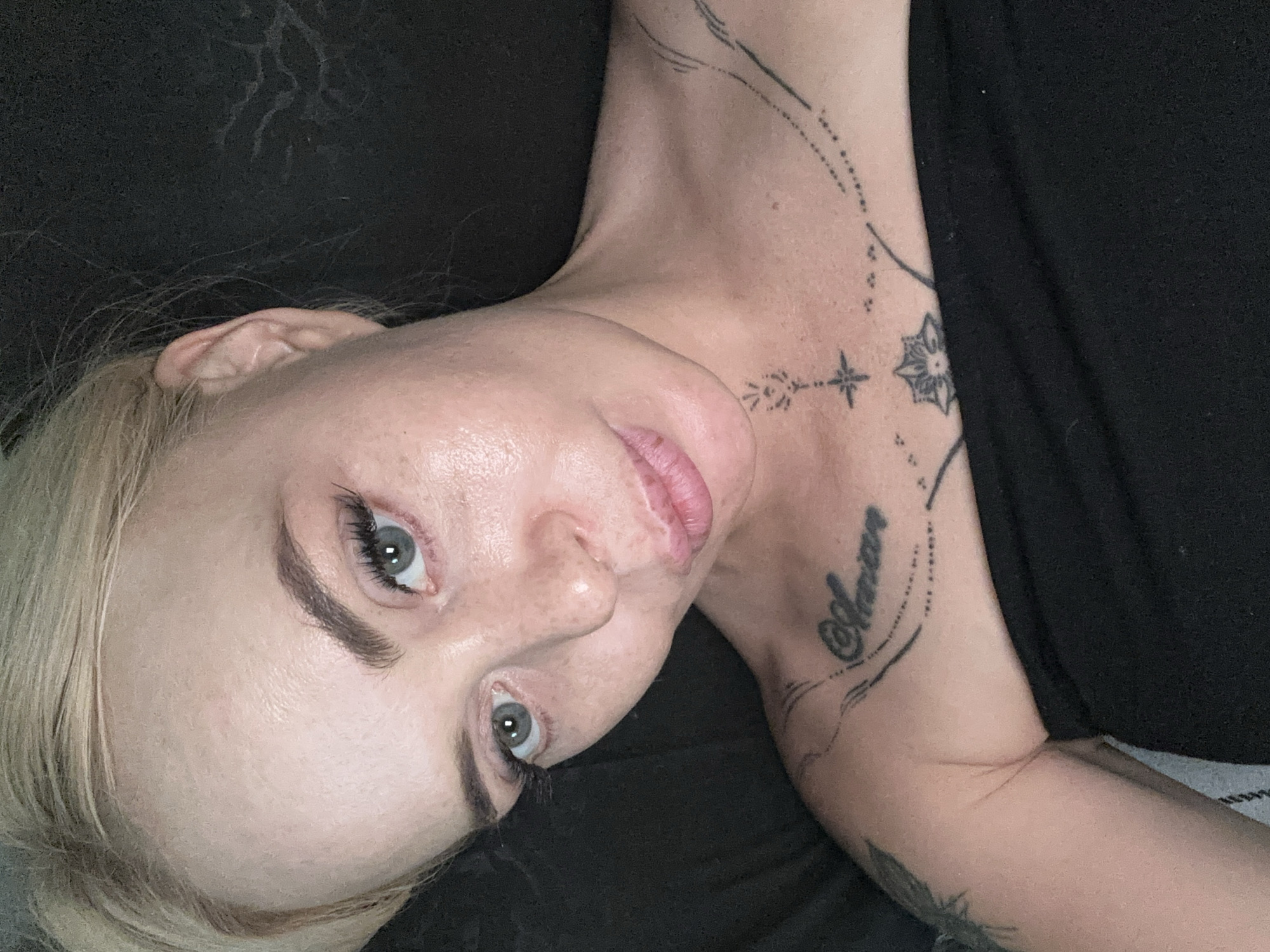 A close-up, high-angle, indoor portrait shows a blonde woman with blue-green eyes, fair skin, and light makeup. She is lying down, with her head tilted and facing the camera. Her hair is blonde and pulled back from her face. She is wearing a strapless black top, and has tattoos on her chest and shoulder. The background is dark and appears to be a bed or pillow. The image has a soft focus and appears to be taken with a flash.