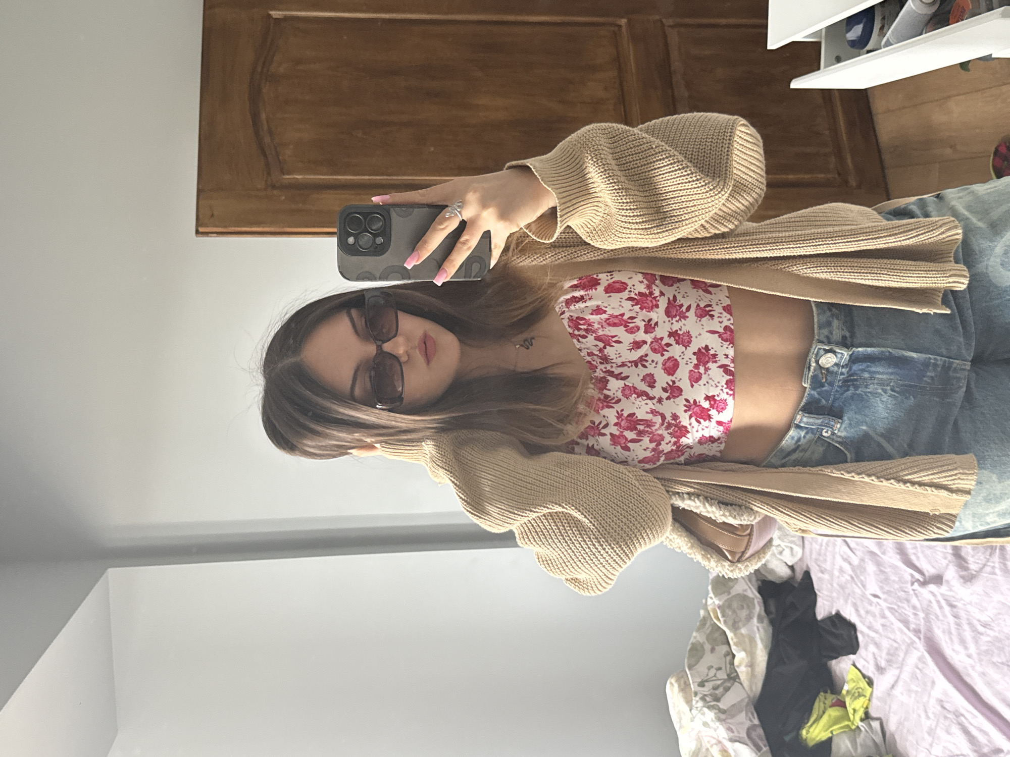 A person is taking a selfie in front of a mirror. The person is wearing sunglasses, a floral top, a beige cardigan, and blue denim shorts. The person is holding a smartphone with a black case. The mirror reflects a wooden door, a white dresser, and a bed with a messy appearance. The location cannot be determined from the image.
