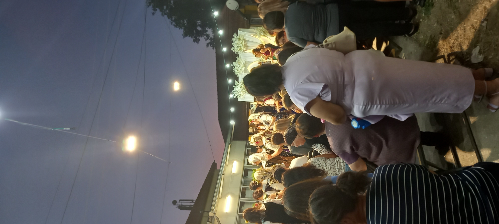 The image depicts an outdoor gathering, likely a celebration or event, taking place at dusk in the city of Lyuben, Bulgaria. In the foreground, several individuals are visible from the back, with one woman in a lavender dress prominently featured. The midground shows a crowd of people, many of whom are wearing traditional attire, including headscarves and folk costumes. Some individuals appear to be dancing or performing. In the background, there is a building with awnings and lighting, suggesting a street or courtyard setting. A white floral archway is visible, indicating a decorated area, possibly for a wedding or a similar ceremony. The sky is a deep blue with hints of purple and orange from the setting sun, and string lights are illuminating the scene. The ground appears to be paved, possibly with concrete or stone. The overall atmosphere suggests a communal and festive event.