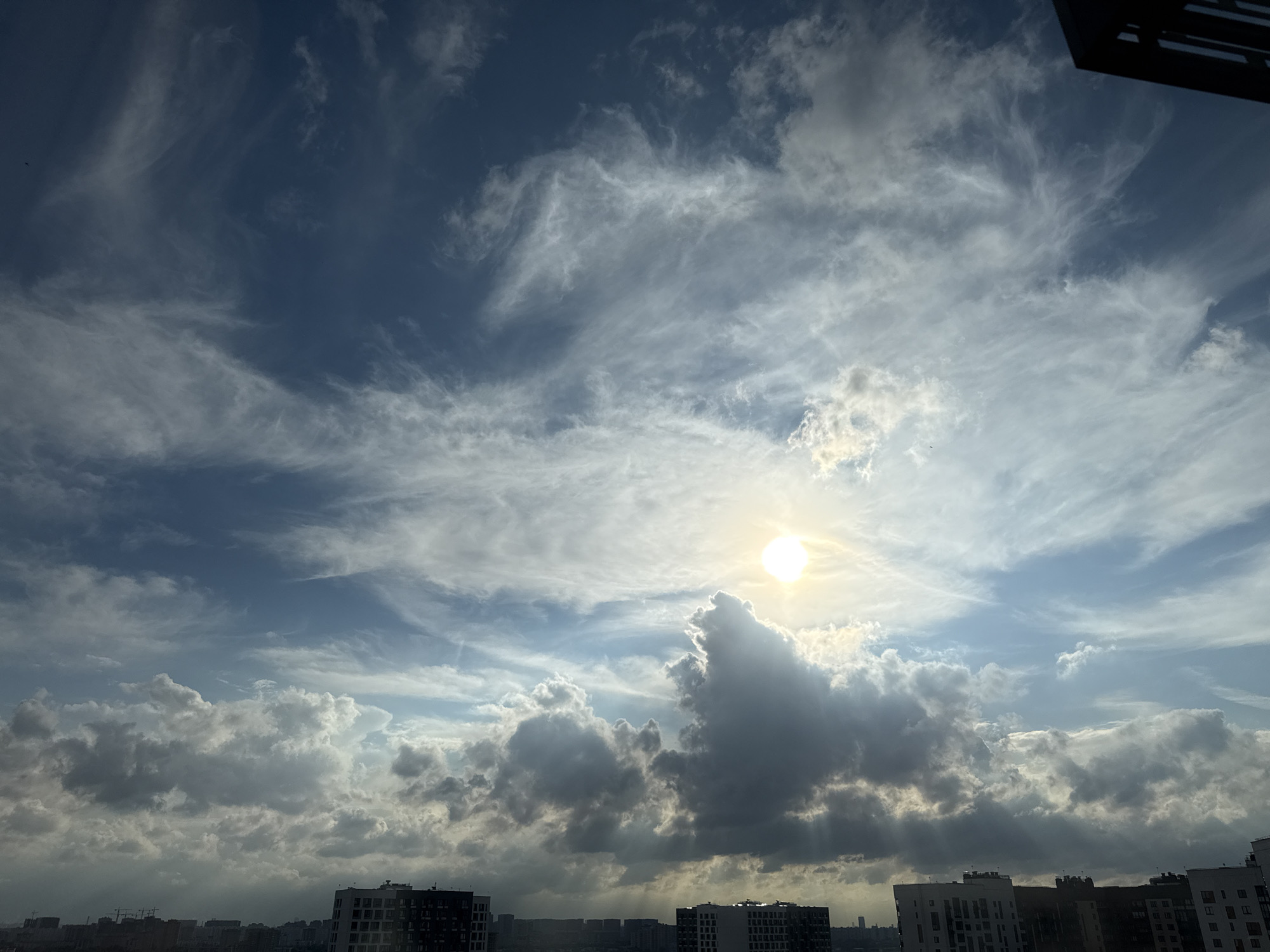 A wide-angle, eye-level shot captures a dynamic sky above the city of Lyubertsy, Russia. The sun is a bright, white orb in the upper right quadrant, partially obscured by dramatic, dark grey clouds that create a sense of depth and shadow. Wispy, white cirrus clouds stretch across the blue sky, adding texture and movement. In the foreground, the rooftops of several apartment buildings are visible, silhouetted against the bright sky. The buildings are modern, with dark and white facades. The overall scene is one of a city bathed in the dramatic light of a partly cloudy day.