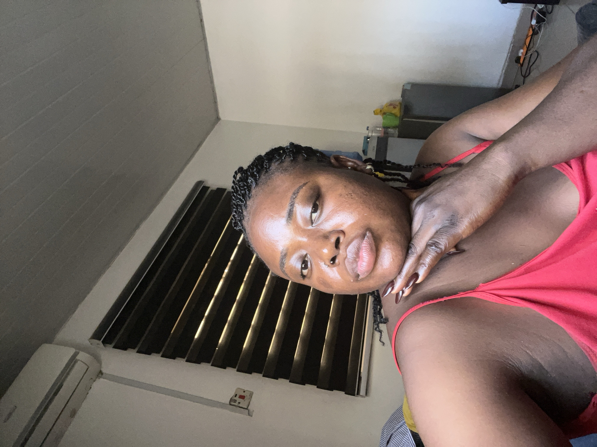 The image is a close-up, slightly angled portrait of a young Black woman indoors. She is lying down, with her head tilted and her hand resting on her neck. Her eyes are partially closed, and her mouth is slightly open, suggesting a relaxed or contemplative mood. She has dark braided hair with some strands loose around her face. Her skin appears to have a dewy sheen, possibly from sweat or skincare. She is wearing a bright red top.
The background includes a window with dark horizontal blinds that are partially open, allowing light to filter through. To the left of the window, there is a white air conditioning unit mounted on the wall. The wall itself is painted a light, neutral color. In the upper right corner, a grey cabinet or shelf is visible, with some items on it including what appears to be a colorful object, possibly a toy or a packet, and a water bottle. Electrical wires and a power strip are also visible connected to something on the shelf.
The lighting suggests it is daytime, with natural light coming from the window. The overall setting appears to be a bedroom or a similar personal space. There is no visible text in the image. The orientation of the photograph is on its side.