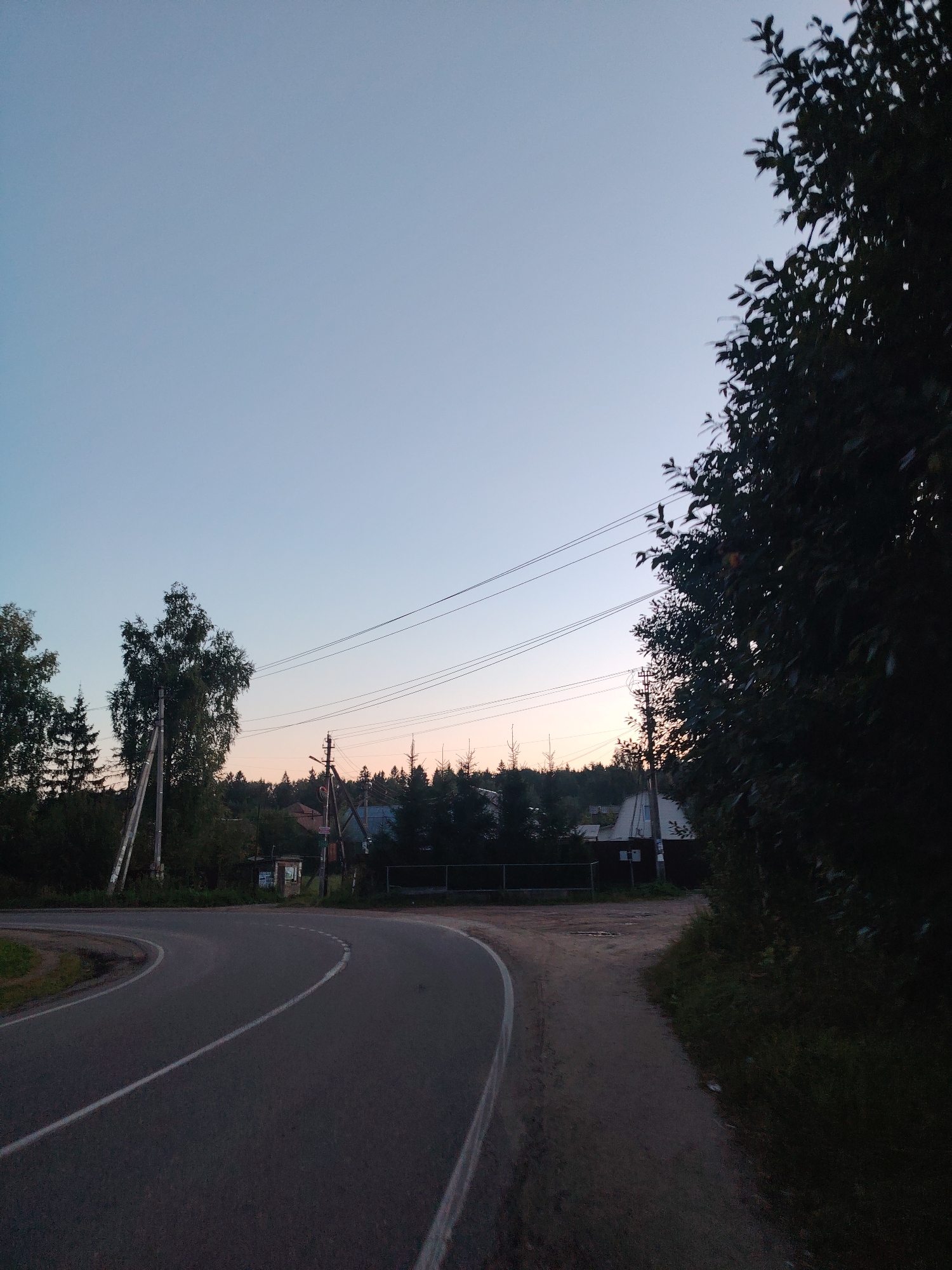 The image depicts a rural scene at dusk in the village of Polyana Malaya SNT, Russia. A paved road curves through the foreground, with a dirt path running alongside it. Several power lines stretch across the sky, supported by wooden poles. In the background, a cluster of houses and trees are visible, nestled amongst a dense forest. The sky is a gradient of pale blue to soft pink, indicating the sunset. There are no people or animals visible in the image. The overall atmosphere is tranquil and serene.
