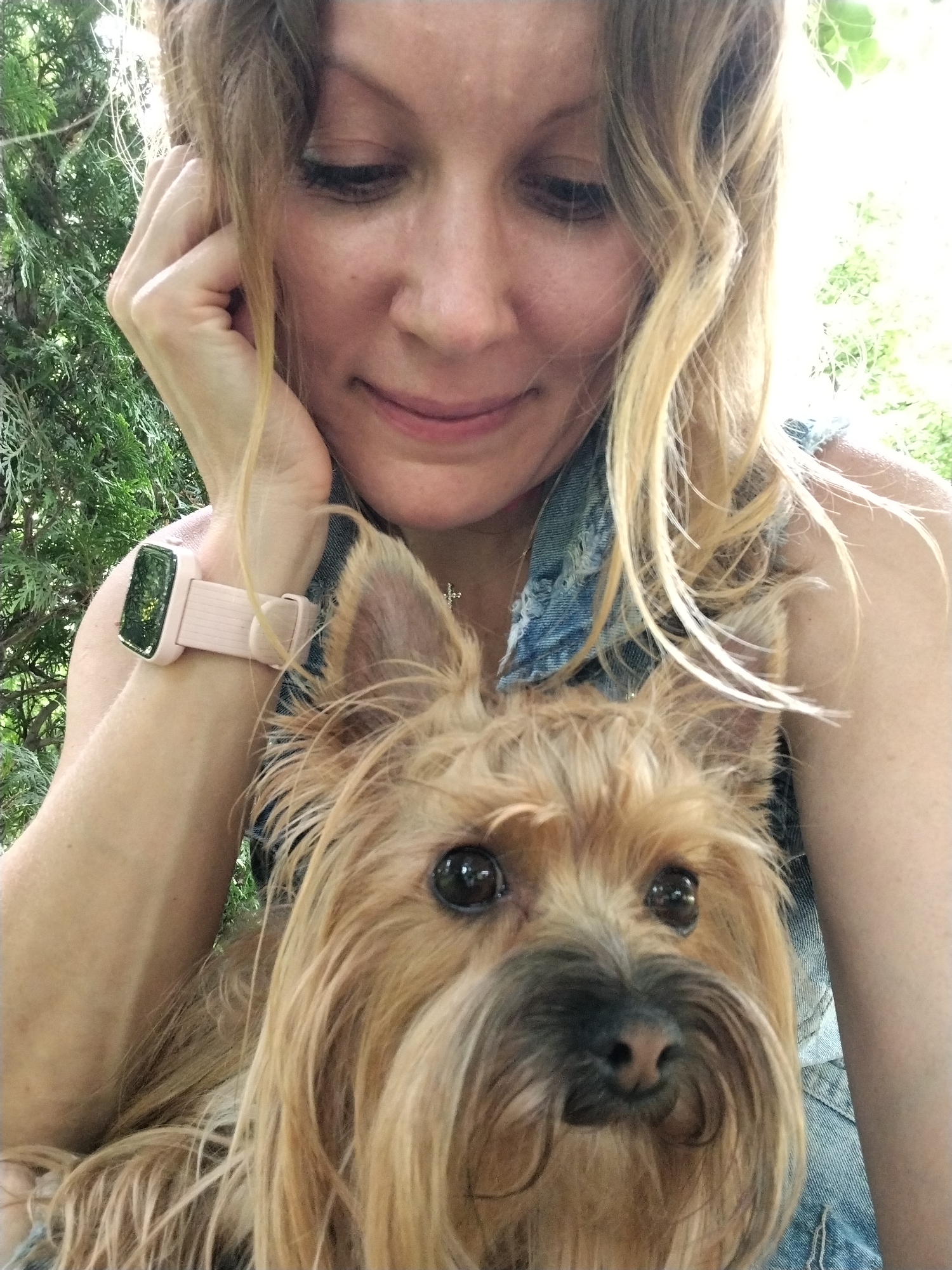 The image is a close-up selfie of a blonde woman holding a small, tan dog. The woman is looking down and to the left, with her left hand touching her face. She is wearing a light pink smartwatch on her left wrist and a denim shirt. The dog, a Yorkshire Terrier or similar breed, is held close to her chest, facing the camera. The background appears to be an outdoor setting with lush green foliage and dappled sunlight. There is no identifiable information within the image to suggest a specific location in Krasnodar, Russia.