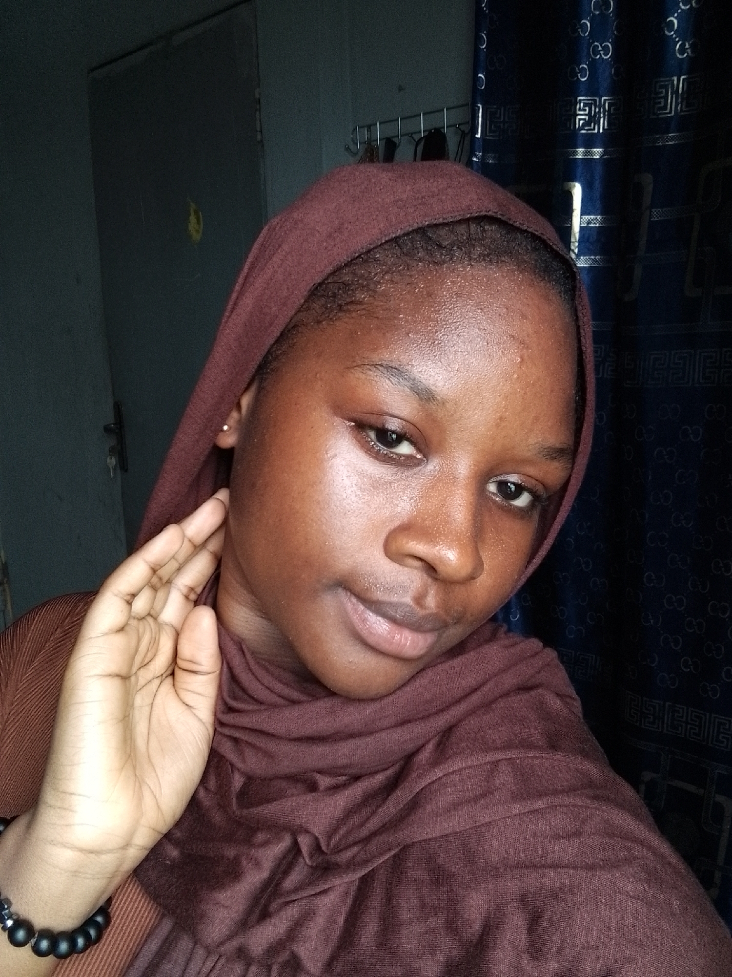 A young woman with dark skin and brown eyes is wearing a brown headscarf. Her left hand is near her face, with a bracelet visible on her wrist. The background features a gray door and blue patterned curtains. Location: Unknown, Unknown.