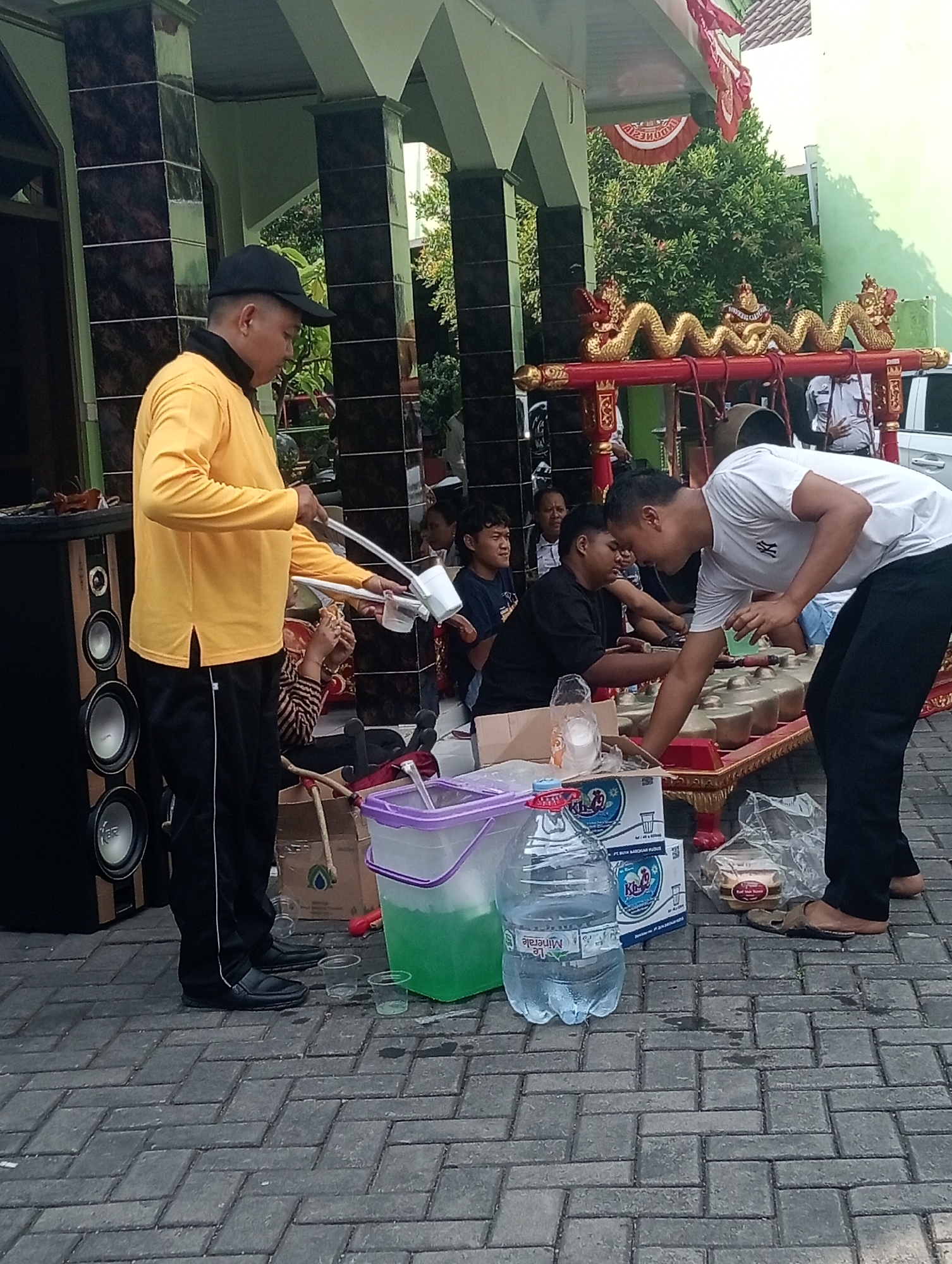 The image depicts an outdoor scene in what appears to be the city of Demak, Indonesia, likely a cultural or community gathering. In the foreground, two young men are engaged in an activity, possibly serving or preparing drinks. One man, wearing a yellow long-sleeved shirt and black pants with a black baseball cap, stands to the left and holds a white pouring tool. The other man, in a white t-shirt and black pants, is leaning over a red and gold decorative structure resembling a traditional musical instrument (likely a gamelan set) and is interacting with small green cups.
Scattered on the ground around them are various items, including clear plastic cups, a large purple-lidded container filled with green liquid, a large bottle of drinking water, several boxes of bottled drinks, and a plastic-wrapped package that appears to be food. To the left, a large speaker system is visible. In the background, several other people are seated, observing the scene. The architectural elements suggest a building with a green facade and dark tiled pillars, possibly a mosque or community hall. Decorative red and gold dragon motifs adorn the structure in the center, hinting at a festive or ceremonial occasion. The ground is paved with interlocking grey bricks. The overall atmosphere appears to be informal and communal.