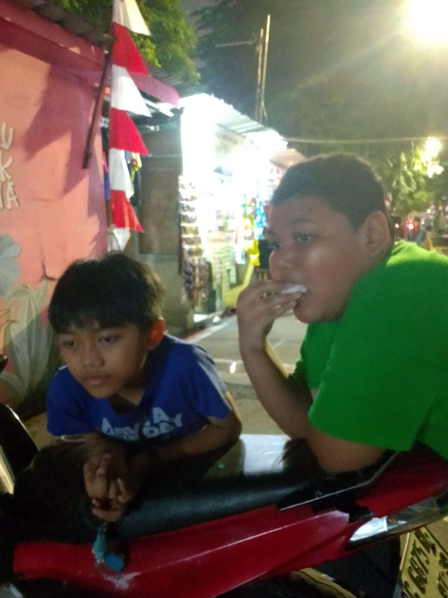 Two young boys are seated on a red motorcycle at night. The boy on the left, wearing a blue shirt, has dark hair and is looking down. The boy on the right, wearing a green shirt, is eating something white and looking to his left. In the background, there is a pink wall with red and white pennants hanging, and stalls with bright lights are visible. The setting appears to be a street market in East Jakarta, Indonesia.