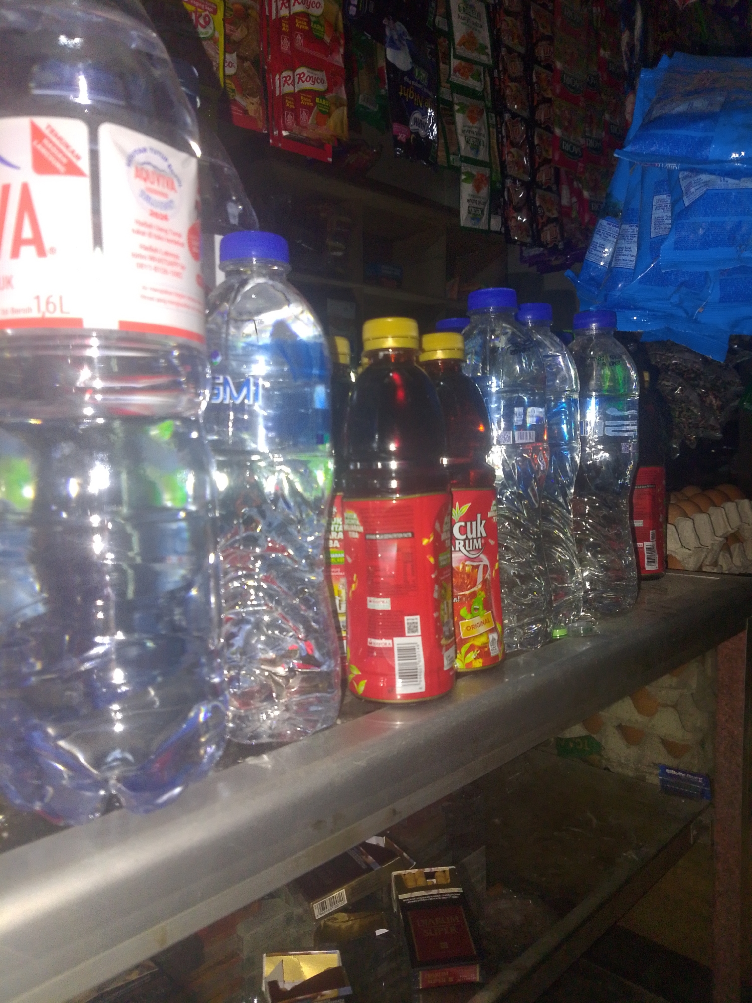 A close-up, slightly blurry, indoor photograph shows a shelf in a small shop, likely a warung, in Mukapayung, Indonesia. The shelf displays a variety of items, predominantly bottled drinks and packaged goods. On the left is a large, clear plastic bottle of water, partially obscured, with a white label featuring red lettering and the "AQUA" logo. To its right are several smaller, clear plastic bottles of water with blue caps. Next to these are two bottles of what appear to be dark liquid, possibly tea or juice, with red labels and yellow caps. Further to the right are more clear water bottles. In the background, on upper shelves, are rows of colorful packets of food seasonings or instant noodles, many with red and white packaging, and blue plastic bags of stacked items. Below the main shelf, partially visible, are cigarette packs and other miscellaneous items. The overall impression is of a typical retail display in a local convenience store.