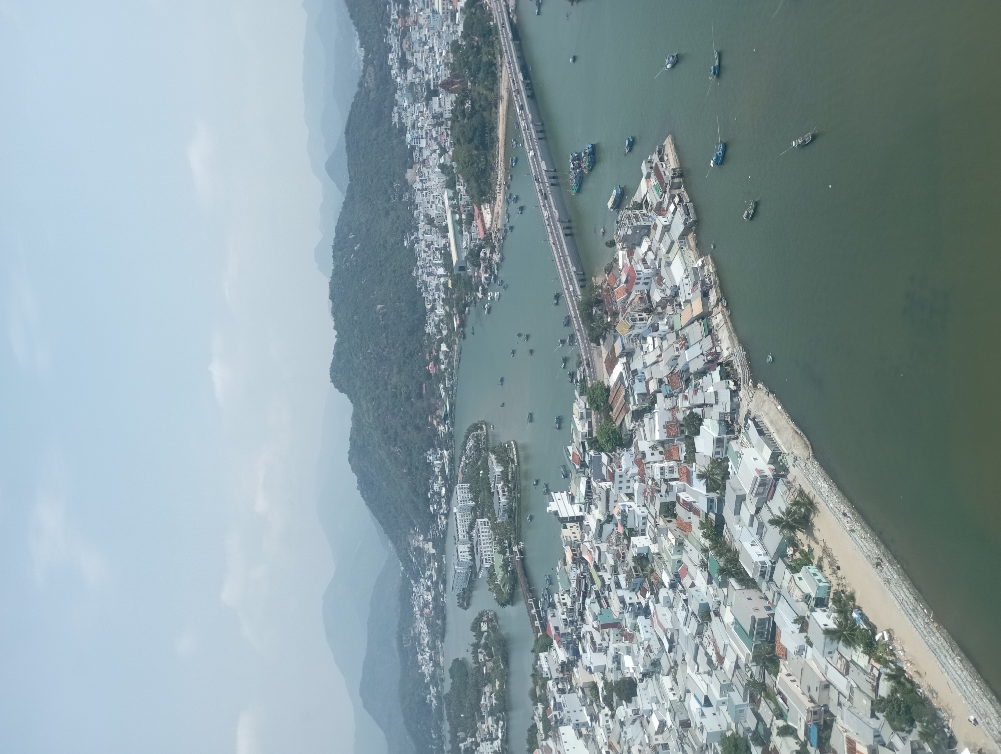 An aerial view shows Nha Trang Ward, Vietnam, a coastal city divided by a river with several bridges. Buildings, largely white and tightly packed, dominate the right side of the image. The coastline is visible, with a rocky breakwater. The river is interspersed with boats and several small islands with buildings and vegetation. A mountain range is seen on the left side of the river, gradually fading into the sky. Bridges span the river, and traffic is visible on the bridges.