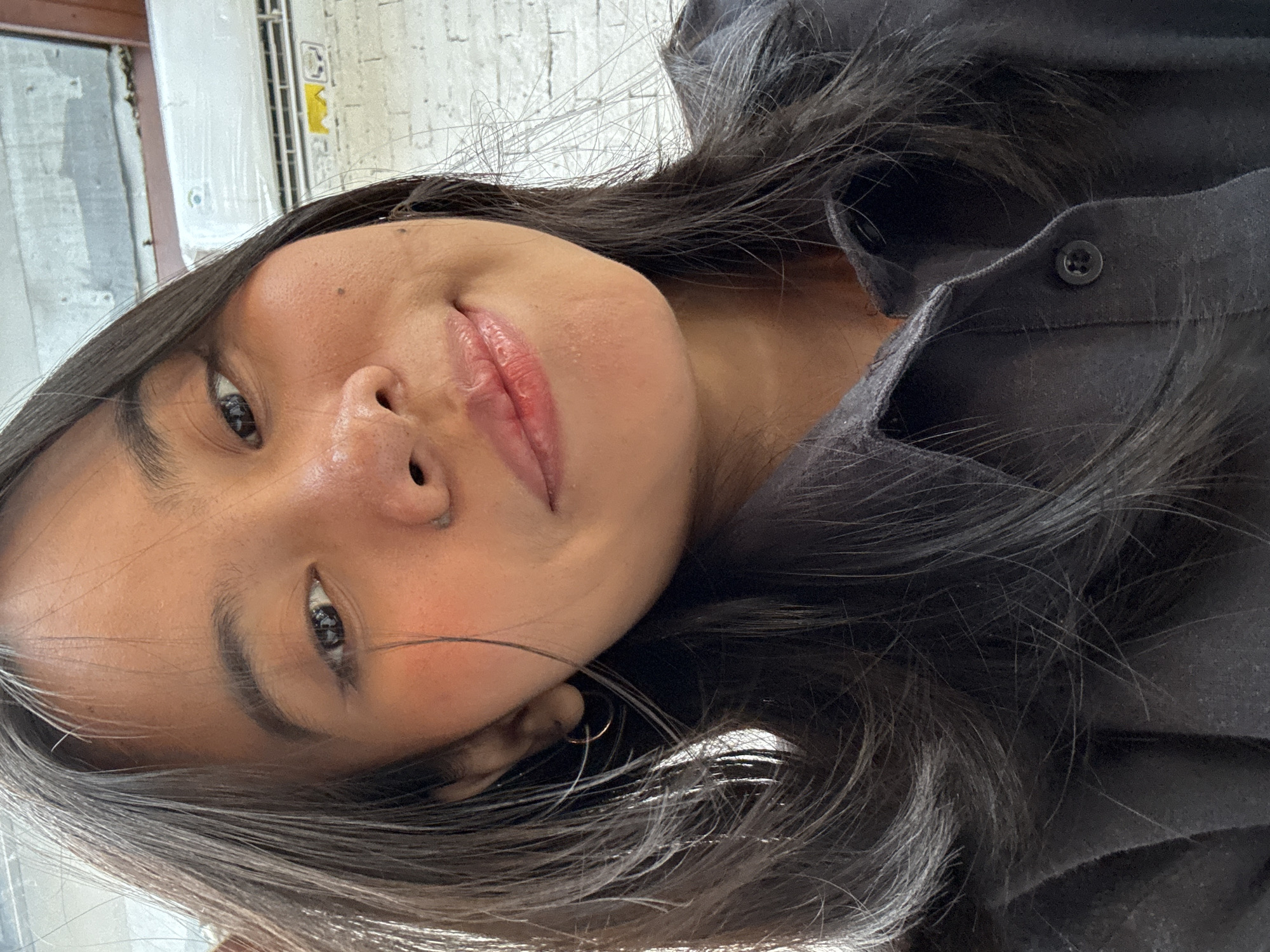A close-up, head-and-shoulders selfie shows a young woman with dark hair, brown eyes, and fair skin, smiling faintly. She is wearing a dark collared shirt. A portion of an air conditioning unit and a white brick wall are visible in the background. The image was taken in South Jakarta, Indonesia.