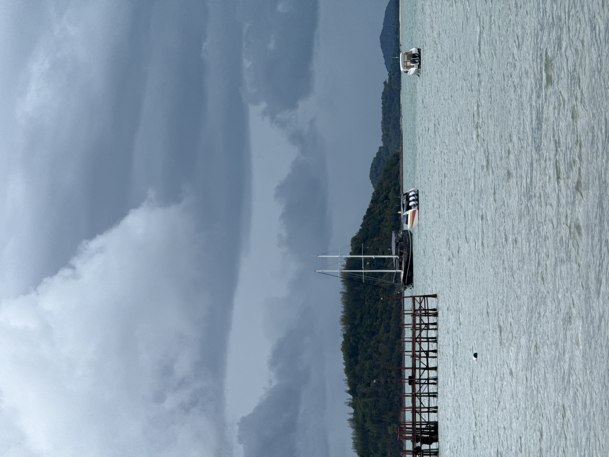 The image depicts a cloudy day at sea in Ko Samui District, Thailand. In the foreground, a wooden pier extends into the choppy, grey water. Several boats are visible in the midground and background. A large, dark sailing vessel with white sails stands to the right of the pier. Two smaller speedboats are also present: one with orange and white markings to the left of the sailing vessel, and another white speedboat further to the right. In the background, a lush green, tree-covered island is visible. The sky is filled with heavy, dark grey clouds, suggesting overcast weather or an approaching storm.
