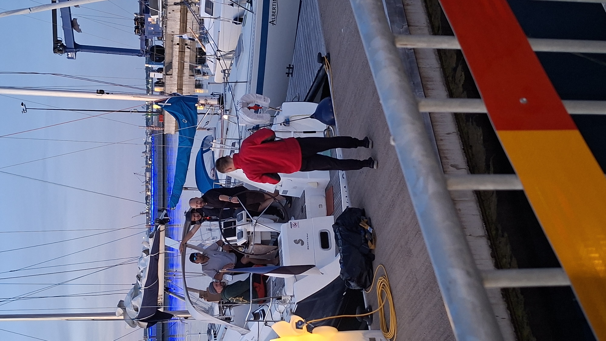 A group of five men are gathered on a dock next to several sailboats. In the foreground, a man wearing a red jacket and black pants stands with his back to the camera, facing a sailboat. He is holding a dark object in his hands. On the sailboat's deck, four men are visible. One man in a grey shirt and black shorts is at the helm, steering the boat. Another man in a black shirt is standing next to him, and two other men are also on the deck. The boats are moored at a dock with a metal railing in the foreground. The sky is a pale blue, suggesting it is either early morning or late evening. The background shows a harbor with cranes and industrial structures, illuminated by blue lights.