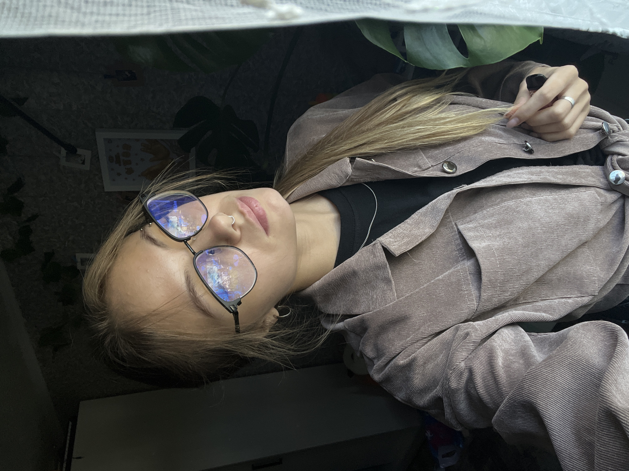A young woman with blonde hair tied in a ponytail is wearing glasses, a black t-shirt, and a brown corduroy jacket. She has a nose ring and is wearing a ring on her finger. The woman is indoors, in what appears to be a room with a window. A large green leaf from a plant is visible on the right side of the image, partially obscuring the woman's face. The background is dark, with a framed picture on the wall and a closet on the left. There are no specific details to indicate the location within the city of Фоки, Russia.