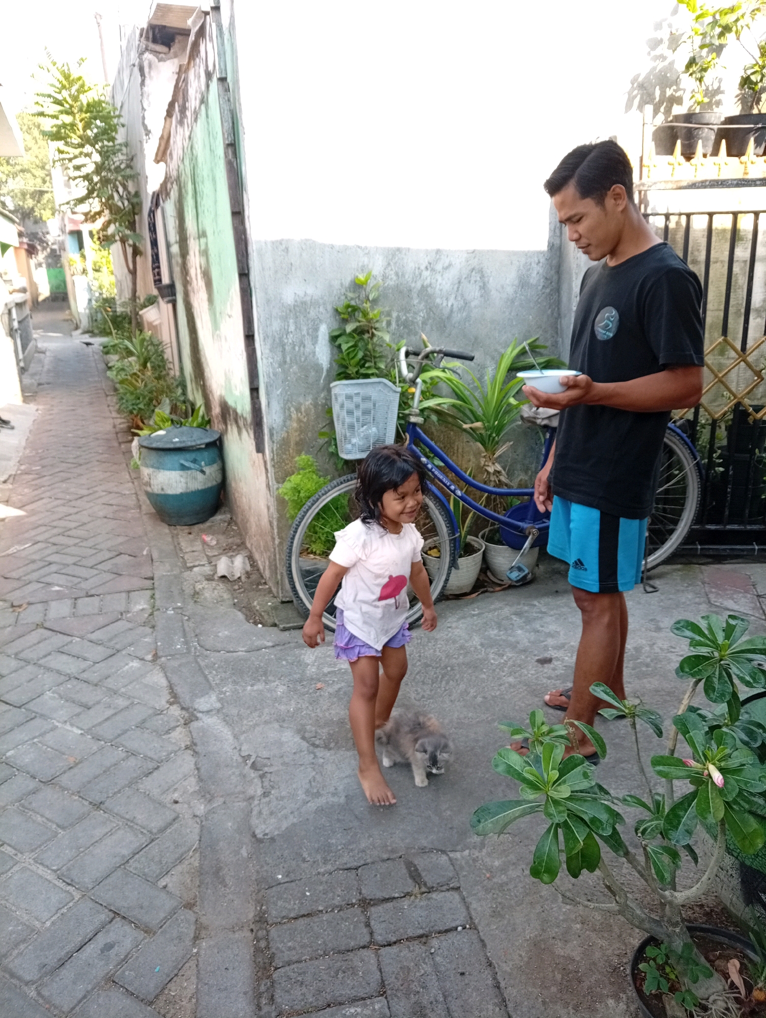 In Surabaya, Indonesia, a man stands near a blue bicycle, holding a white bowl. A young girl in a pink shirt and purple shorts stands before him, looking down at a small grey cat on the cobblestone ground. The environment appears to be a narrow alleyway with a concrete wall on one side and a potted plant in the foreground.