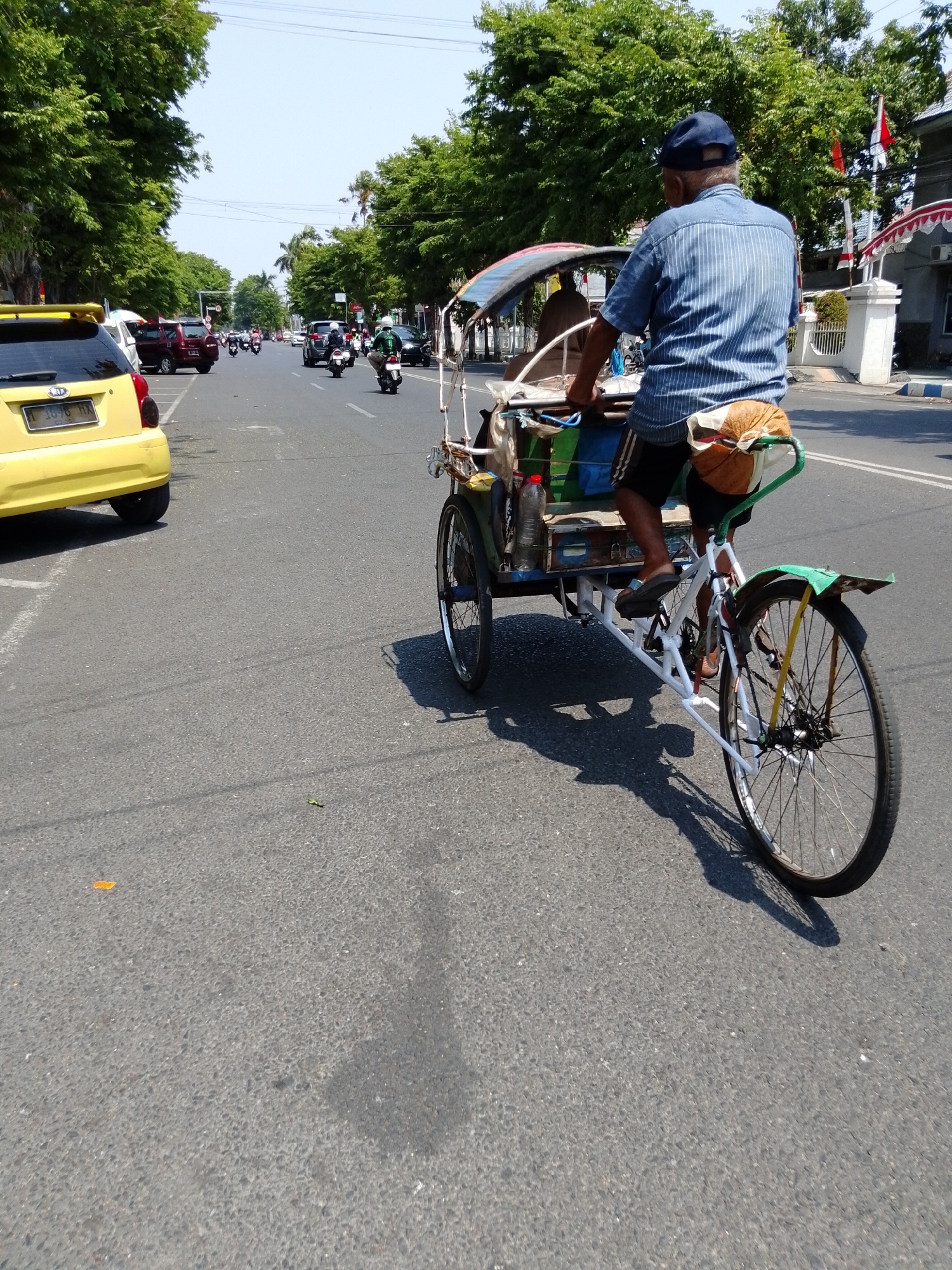 The image depicts a street scene in Pasuruan, Indonesia, on a bright, sunny day. In the foreground, a three-wheeled bicycle rickshaw is moving along the asphalt road. The rickshaw is white with green accents on the fenders and handlebars. It carries various items in its cargo area, including colorful bags and a plastic water bottle. A man wearing a blue striped shirt, dark shorts, and a dark blue baseball cap is pedaling the rickshaw, looking over his right shoulder. Behind him, the passenger compartment of the rickshaw is visible, containing a person.
To the left of the rickshaw, a yellow hatchback car is parked. The car has a license plate that reads "L 1696 HX". Further down the street, other vehicles, including cars and motorcycles, are visible. The road is lined with lush green trees on both sides, and the sky is clear blue. In the background, buildings with red and white awnings can be seen, suggesting a commercial or residential area. The overall impression is of a typical day in a moderately busy Indonesian city.