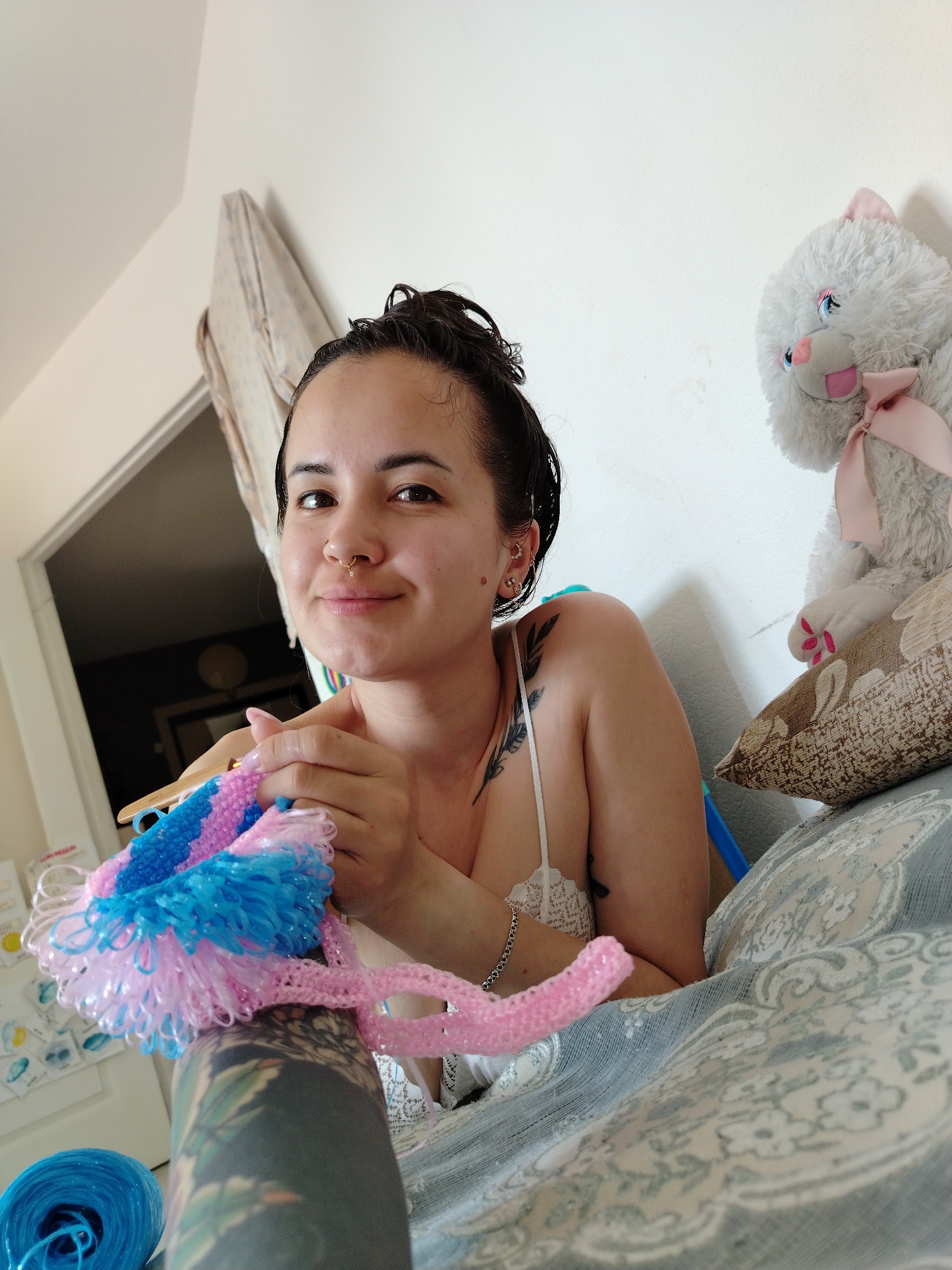 A person with short, dark, damp hair, multiple ear piercings, and a nose ring is crocheting with blue and pink yarn. The person has tattoos on their right arm and shoulder. They are lying on a patterned bed, with a stuffed cat propped up on pillows next to them. A blue ball of yarn is on the bed near the person. The location is a room with white walls and a doorway visible in the background. The city of Noviy, Russia is not discernible from the image.