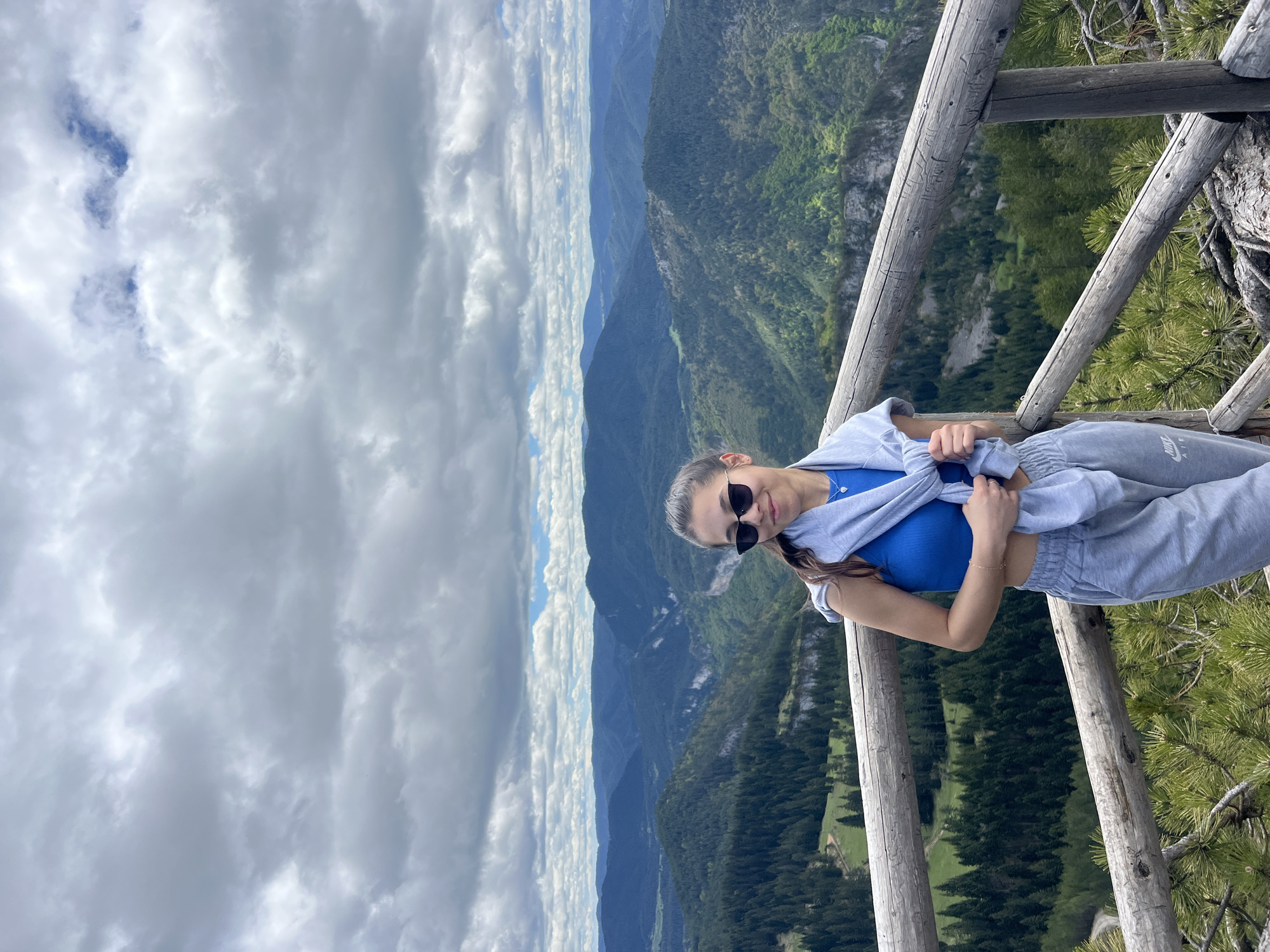 A woman is standing on a wooden observation deck overlooking a mountainous landscape in Giovren, Bulgaria. The woman has sunglasses, a blue top, and a gray tracksuit. She is holding a gray jacket or sweatshirt in her hands. The background consists of layered mountain ranges covered in green vegetation, extending into the distance. The sky above is filled with a mix of white and gray clouds. The wooden deck is constructed with round wooden posts and railings. Some evergreen trees are visible near the railing of the deck.
