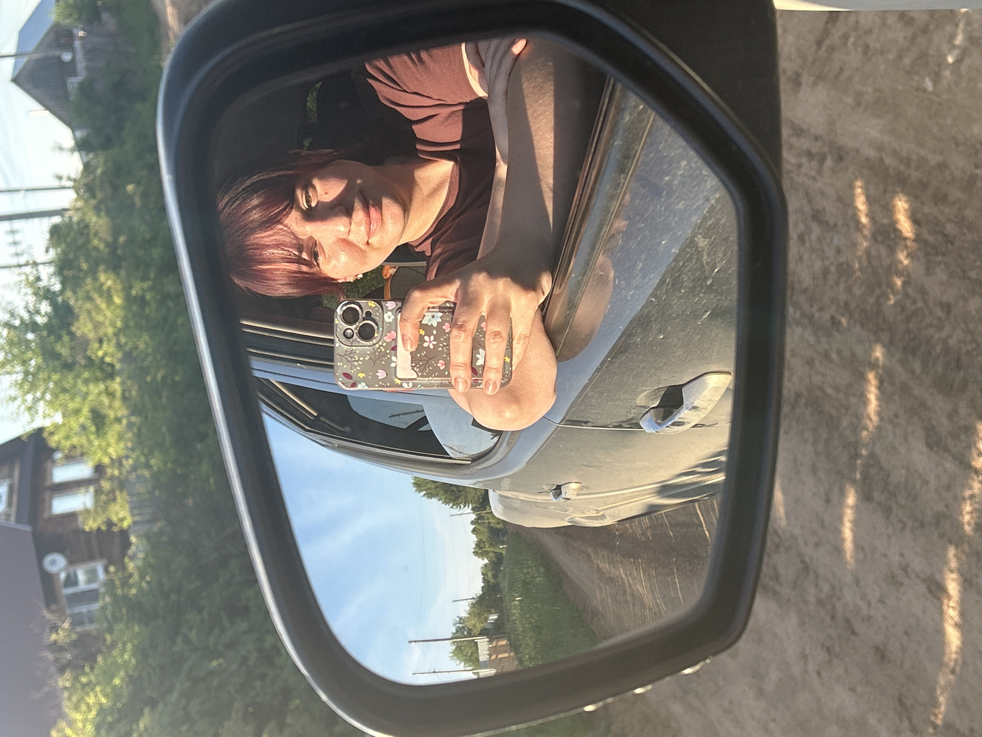 A woman is taking a selfie in a car's side mirror. She has red hair and is wearing a maroon t-shirt. Her left arm is resting on the open car window. The car is driving on a dirt road, with green bushes and trees on either side. In the background, there is a wooden house. The sky is blue with some clouds. The scene appears to be in a rural area.
