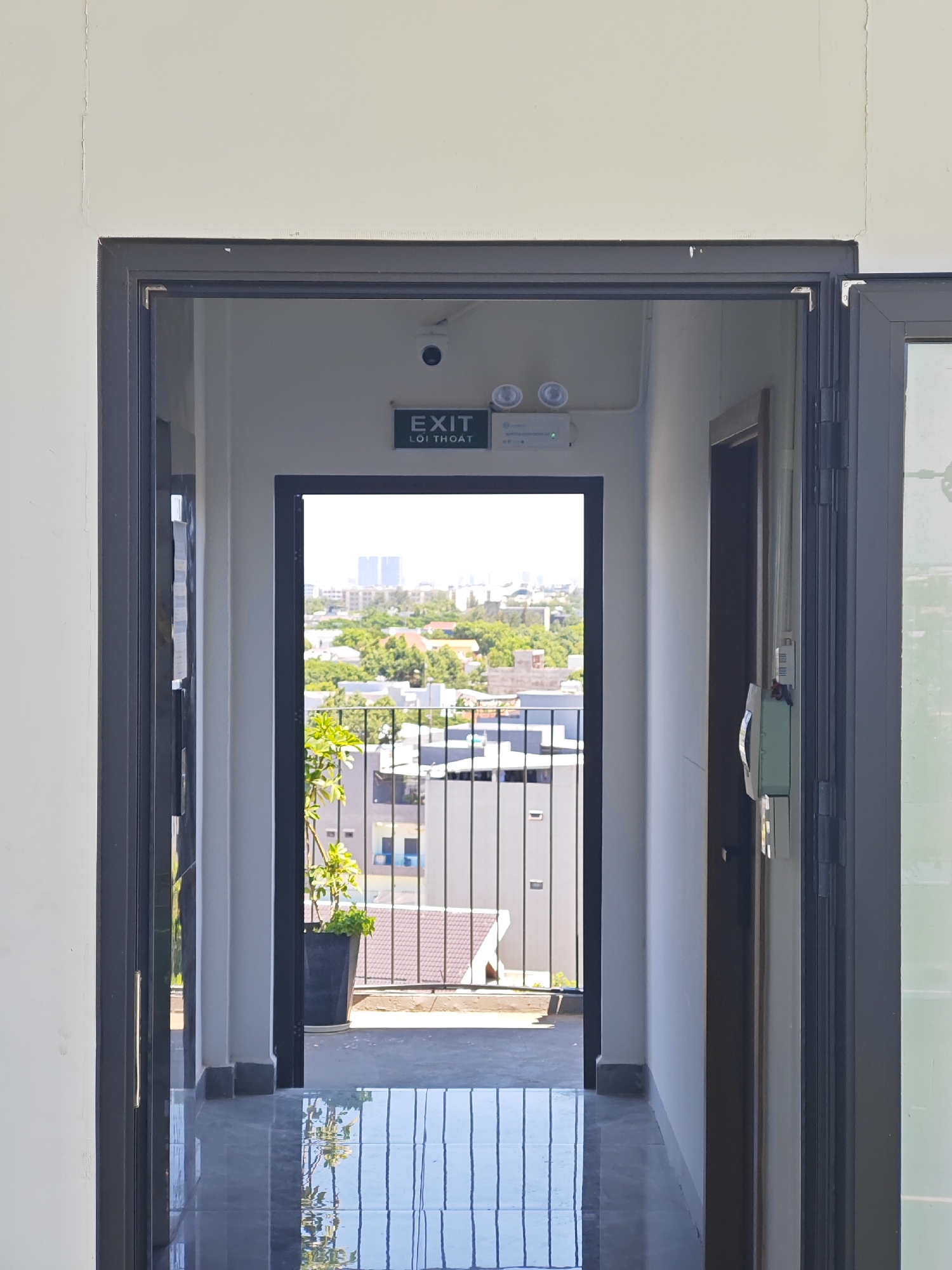 The image captures a view through an open doorway, revealing a hallway that leads to an outdoor balcony or terrace. Inside the hallway, a green illuminated exit sign with Vietnamese text "LÕI THOÁT" is visible on the wall, accompanied by a security camera and two emergency lights. A potted plant with green leaves stands near the doorway, on the left side of the frame. The hallway flooring is polished and reflects the light. Beyond the doorway, a balcony with a metal railing overlooks a city landscape characterized by low-rise buildings and lush trees, with taller buildings in the distance. The sky is overcast. This scene is located in the city of Đà Nẵng, Vietnam.