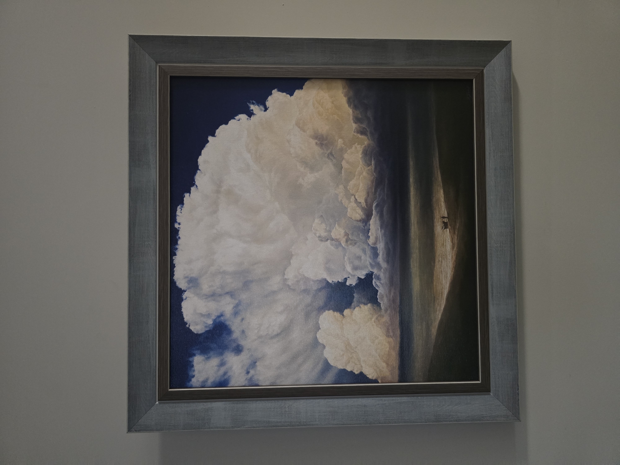 A framed painting hangs on a light gray wall. The painting depicts a large, fluffy white cloud filling the left side of the frame against a dark blue sky. To the right of the cloud, the painting transitions into a vertically oriented landscape with darker, shaded tones giving way to brighter, sunlit areas. Two tiny figures, appearing to be humans, are visible standing in the sunlit area. The painting is enclosed in a gray frame with a subtle, weathered texture.