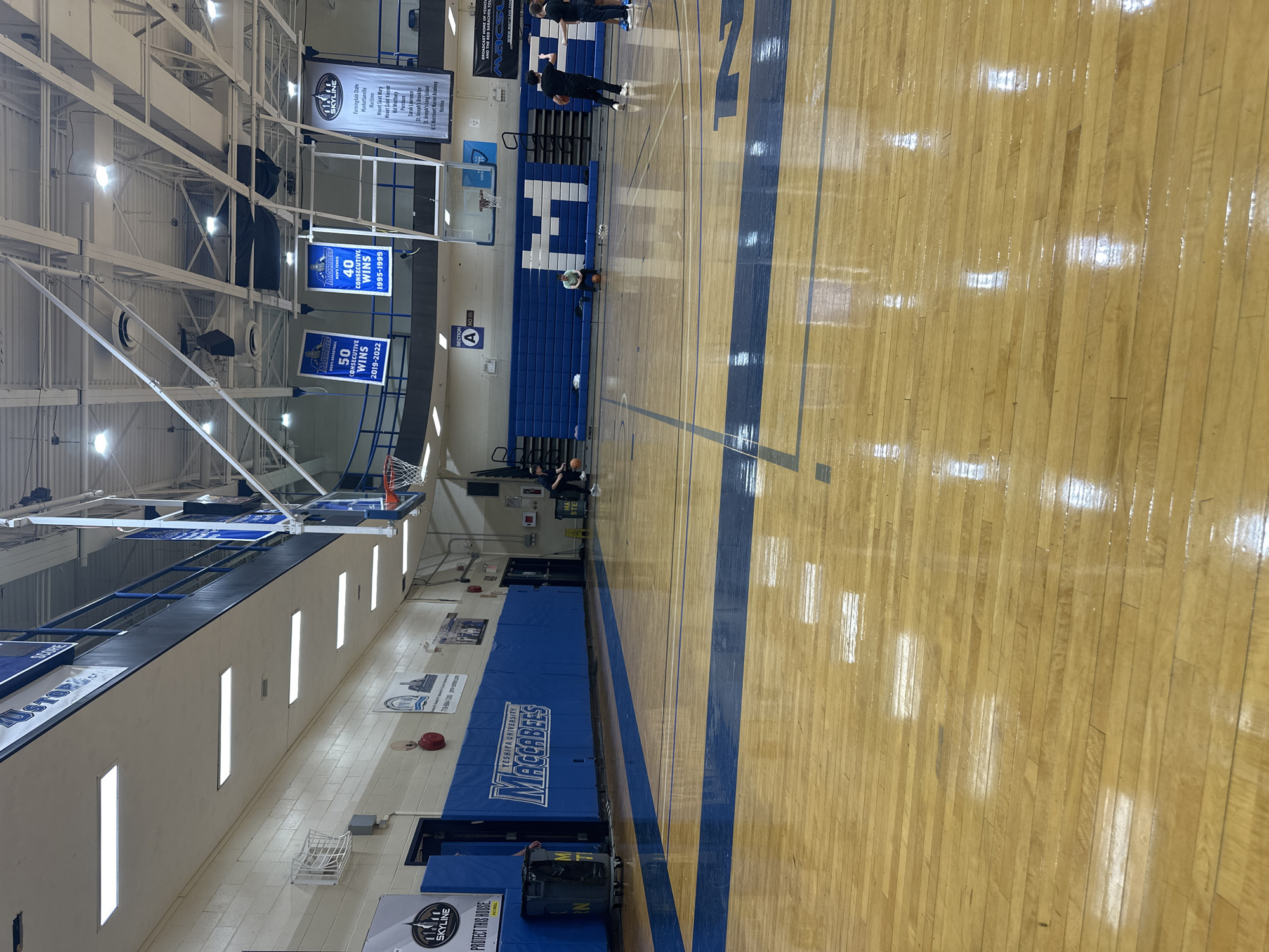 An indoor basketball gymnasium at Yeshiva University in New York City, United States of America, is depicted. The polished hardwood court features blue and black lines, reflecting bright overhead fluorescent and high-bay lighting. The left wall is constructed of light-colored cinder blocks, with a lower section covered in blue padding displaying "YESHIVA UNIVERSITY MACCABEES" in white text. Above this padded section is a blue-railed balcony.
Banners suspended from the ceiling commemorate athletic achievements, including "Maccabees Men's Basketball 50 Consecutive Wins 2019-2022" and "Maccabees Men's Tennis 40 Consecutive Wins 1995-1999." Two basketball hoops with glass backboards and white nets are visible. Blue retractable bleachers are positioned against the far wall, displaying a large white "M" logo. A yellow "WET FLOOR" sign is present near the bleachers.
Four individuals are on the court: one person in black attire actively dribbles a basketball in the mid-right area; a second person, also in black, stands adjacent; a third person, wearing a light green shirt, sits on a basketball near the far wall; and a fourth person, partially obscured, stands near a black trash receptacle on the left side of the court.