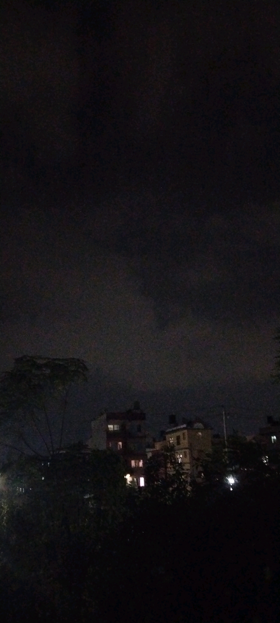 The image captures a nighttime scene in Yama, Nepal. The sky is dark and cloudy, with some subtle variations in tone suggesting the presence of diffuse light. In the middle ground, several multi-story buildings are visible, their architecture indicative of urban construction in the region. Some windows in these buildings are illuminated, indicating occupancy. In the foreground, dark silhouettes of trees and foliage obscure parts of the lower section of the image. The overall impression is one of a quiet, possibly residential area at night.