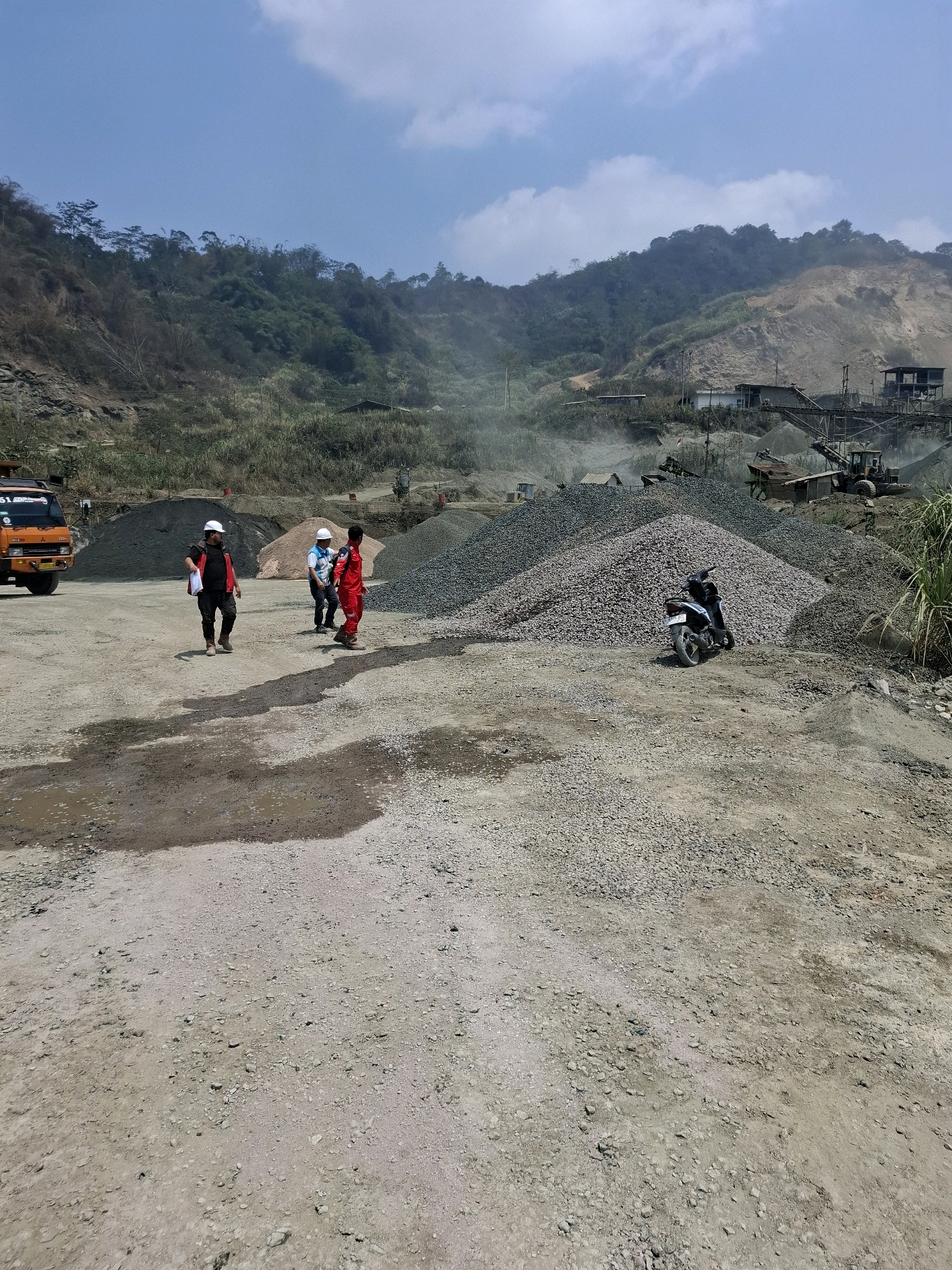 The image depicts a quarry operation in the city of Cijangkar, Indonesia. In the foreground, three individuals, two wearing hard hats and one in a red uniform, are walking on a dusty, gravelly surface. There are puddles of water on the ground. To the left, an orange truck is parked. In the mid-ground, large piles of gravel and stone are visible, along with some industrial equipment and structures, including what appears to be a conveyor belt system and a building. A black motorcycle is parked on one of the gravel piles. The background features a hilly, vegetated landscape with some areas of exposed rock and soil, suggesting ongoing quarrying activities. The sky is clear with some clouds.