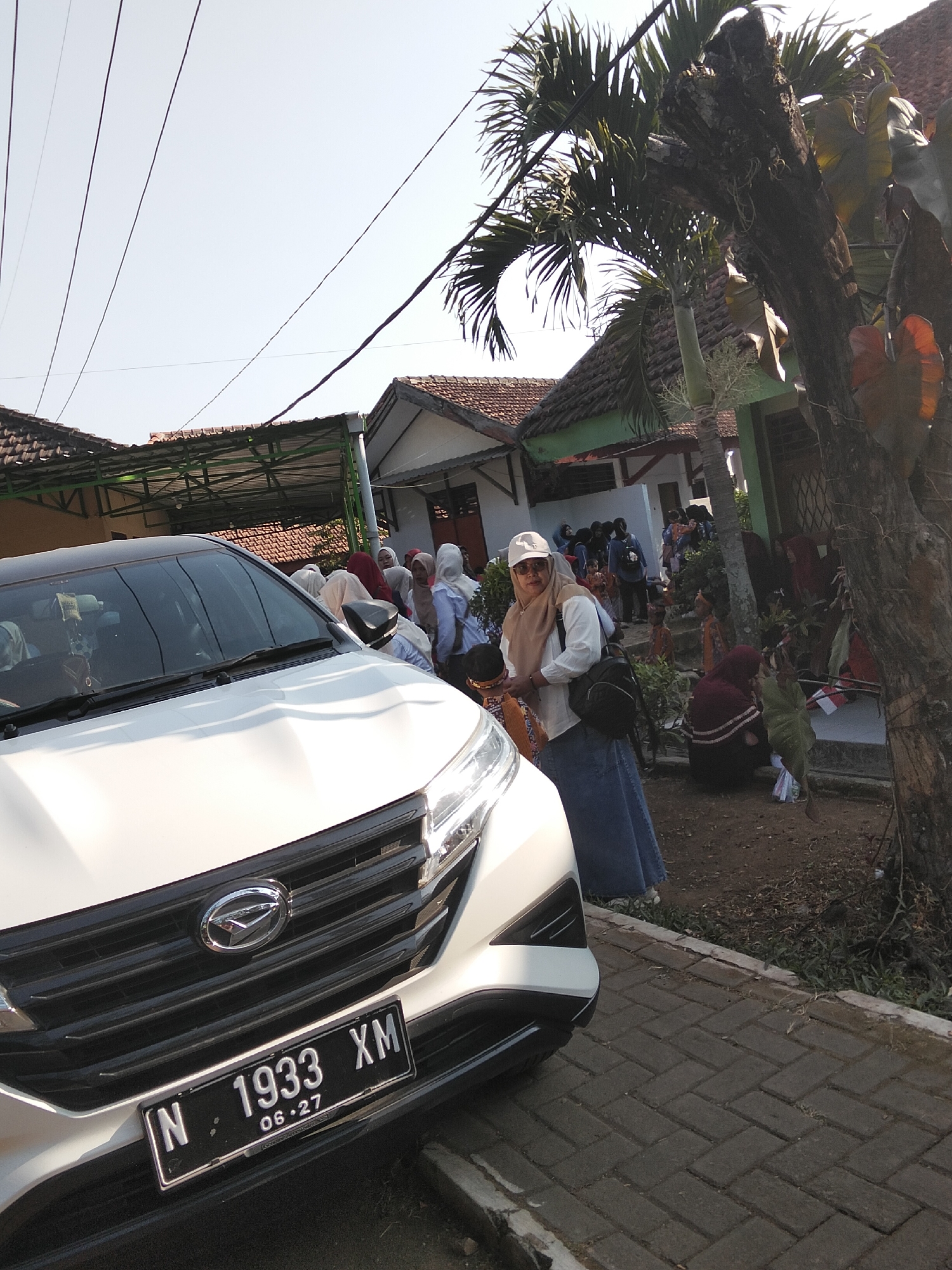 In Puspo, Indonesia, a white SUV is parked on a brick pathway in front of a building with a green trim and tiled roof. A woman wearing a tan headscarf, white shirt, sunglasses, and a blue skirt stands to the left of the vehicle, facing the camera. She is holding a black backpack. Behind her and the car, a group of people, mostly women wearing headscarves, are gathered. Some are standing, and others are seated on steps leading into the building. A large tree with rough bark is on the right side of the frame, with palm fronds extending from the top. The sky is clear and blue, indicating a sunny day. The car's license plate is visible and reads "N 1933 XM 06-27".