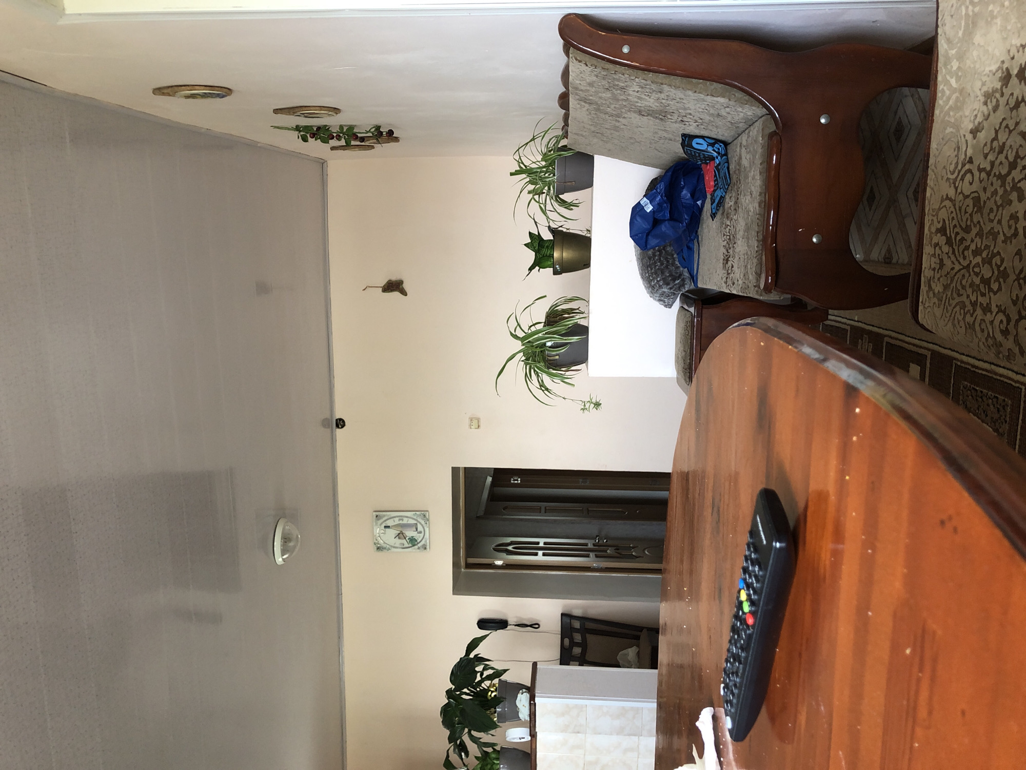 The image shows an interior scene, likely a living room or dining area, with a warm, neutral color palette. In the foreground, a polished wooden dining table occupies the lower left quadrant, with a black remote control with colored buttons resting on its surface. To the right, a wooden bench or sofa with upholstered cushioning and decorative wooden trim is visible. Several potted plants are placed on a white shelf or ledge against the wall. A doorway leading to another part of the house is visible in the background. Decorative elements, including framed pictures and a clock on the wall, add to the domestic setting. The ceiling is a light color, with a visible light fixture. The overall impression is one of a lived-in, comfortable home. There are no people or animals visible in the image.
