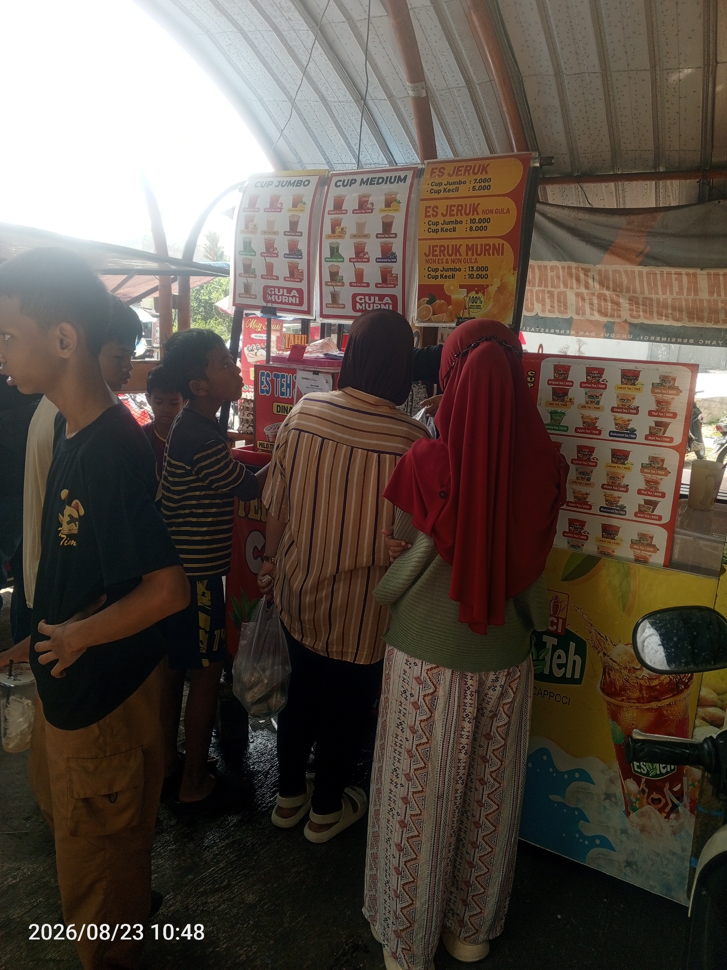 This is a medium shot of a group of people at an outdoor drink stand. The stand has a metal roof and is decorated with colorful signs advertising various drinks, including "ES JERUK" (orange juice) and "ES TEH" (tea). The people appear to be Indonesian. In the foreground, a young person wearing a black t-shirt and tan cargo shorts stands to the left, facing away from the camera. To their right are two younger boys, one wearing a striped t-shirt and the other a dark t-shirt. Behind them, two women are visible, one in a brown and white striped shirt and the other wearing a red hijab. The woman in the red hijab is looking at a menu. The ground is wet, suggesting it may have recently rained. The overall scene depicts a common street scene in Indonesia, likely a local market or roadside stall. The timestamp in the bottom left corner of the image reads "2026/08/23 10:48," which is in the future and suggests this is a simulated date.