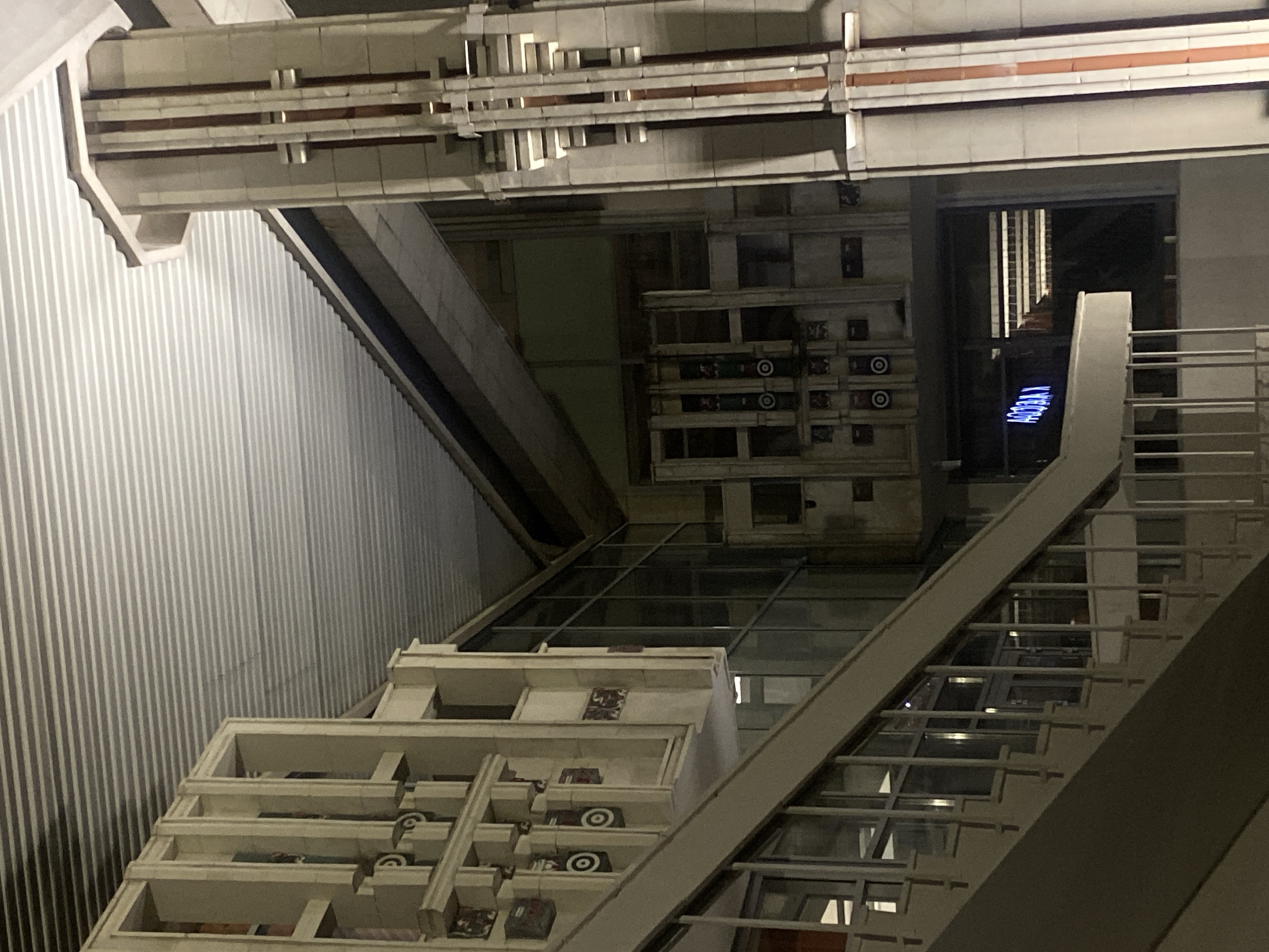 The image depicts the interior of a modern building, likely an atrium or multi-level space, captured at what appears to be evening or night due to the artificial lighting. The perspective is looking upwards, showcasing a complex architectural environment.
On the left side of the frame, a tall, light gray corrugated metal wall provides a textured backdrop. To its right, a prominent, wide staircase or escalator with sturdy railings ascends upwards. Beneath this, a large sloped section composed of multiple glass panels suggests a skylight or a structural glass wall.
In the lower foreground, a multi-tiered display structure is visible, resembling stacked open boxes or shelves. Each compartment holds miniature vehicles, such as classic cars or trucks, and other small artifacts, forming what appears to be an art installation or an elaborate museum exhibit.
Above the main staircase, the building's ceiling or an upper floor features intricate architectural detailing. This structure consists of intersecting beams and panels, creating a geometric pattern with linear elements and varied textures, suggesting concrete or stone.
In the far background, seen through an opening or window, a vivid blue neon sign is visible. Upon close examination and mental rotation, the sign clearly spells "VERTIGO" when read vertically from bottom to top.
The scene is devoid of people, emphasizing the architectural design and the static display. The overall aesthetic is industrial and contemporary, with a strong focus on lines, patterns, and artificial illumination.