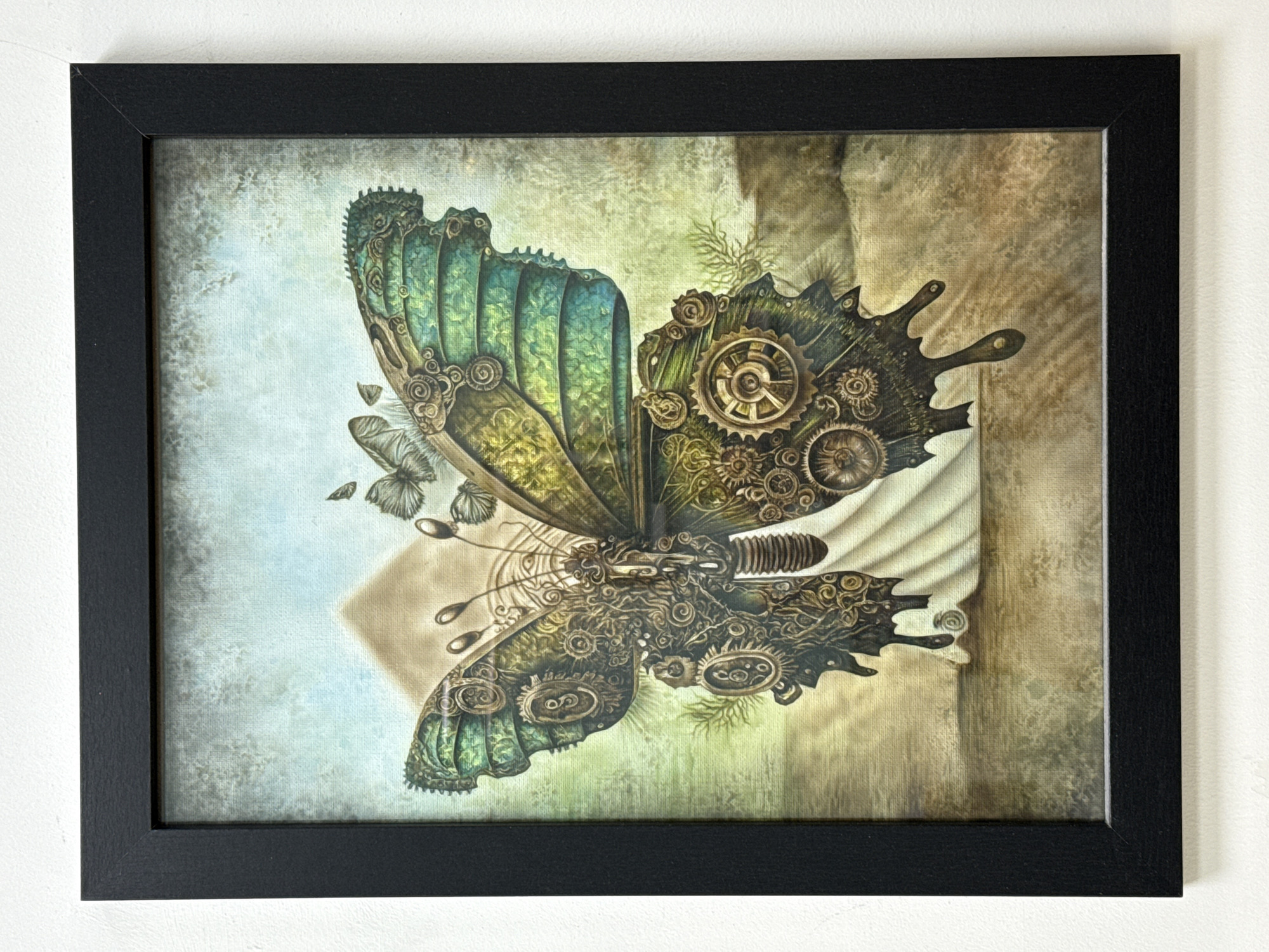 A framed painting hangs on a white wall. The painting depicts a steampunk-style butterfly. The left side of the butterfly is green and blue, while the right side is brown and features gears and mechanical components. The background of the painting is a mix of muted colors, with a diamond shape behind the butterfly. The frame is black. There are no people visible in the image. The location of Burgas, Bulgaria is not apparent in the image.
