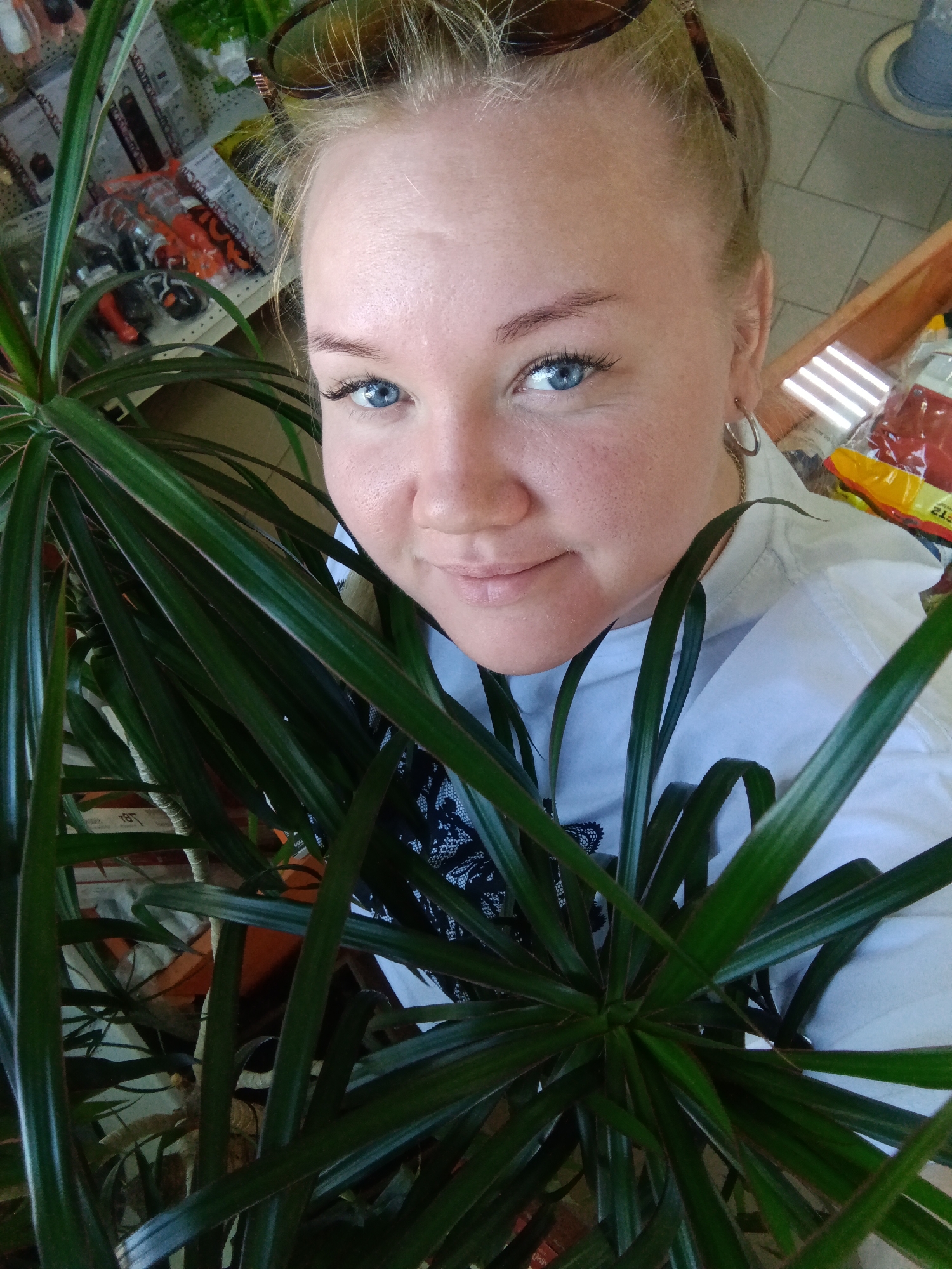 A woman with blonde hair pulled back and blue eyes is posing for a selfie. She is surrounded by a green plant with long, thin leaves. The background appears to be a store, possibly a hardware or garden supply store, with shelves visible behind her. The location might be Фоки, Russia, based on the context.