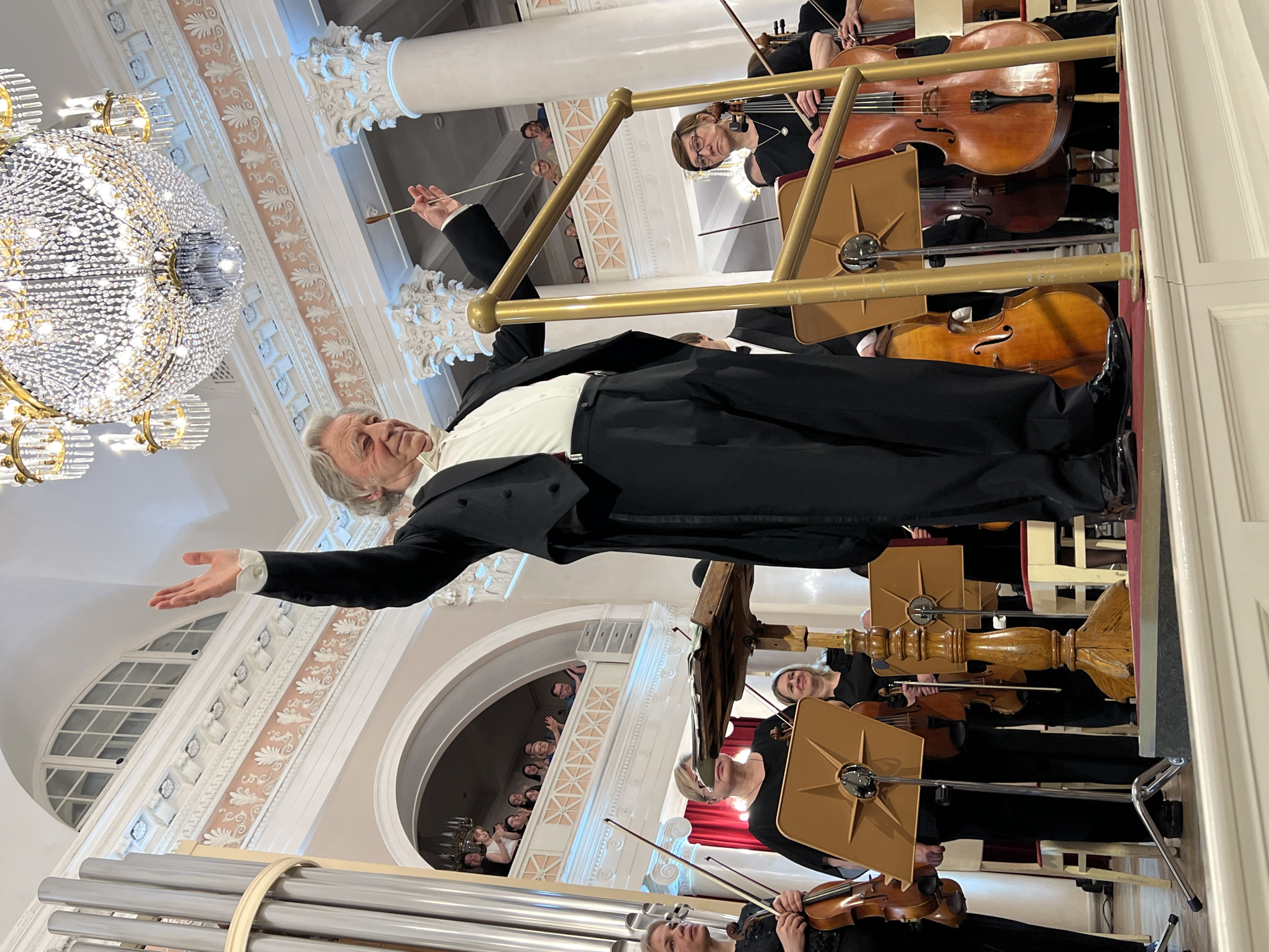 The image depicts a conductor in a black tuxedo with grey hair conducting an orchestra. He is positioned on a stage with musicians playing string instruments such as violins and cellos. There are music stands in front of each musician. A large, ornate chandelier hangs from the ceiling. The venue appears to be an elaborate concert hall with arched windows and architectural details. The hall is populated with people, who appear to be enjoying the performance. The location is likely in Saint Petersburg, Russia, given the architectural style and the cultural context suggested by the orchestra and concert hall.