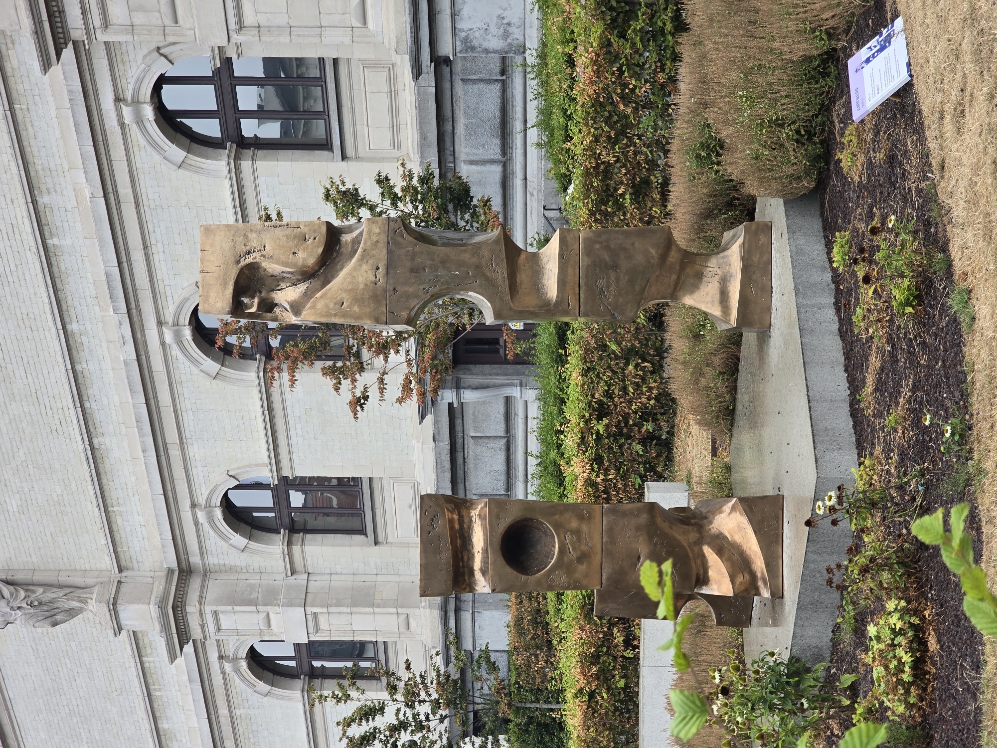 The image shows two abstract bronze sculptures in front of a building with a light-colored brick facade. The sculptures are positioned on a concrete platform, with manicured bushes and dry grass surrounding them. In the foreground, there are some green leaves and small plants. The building features arched windows and ornate architectural details. The overall scene suggests an outdoor art installation in an urban setting.
The sculptures are located in the city of Antwerp, Belgium, specifically at the entrance of the Museum of Fine Arts (KMSKA). The sculpture on the left is a stacked, block-like form with a circular opening in the center. The sculpture on the right is a taller, more organic, and twisted form.
