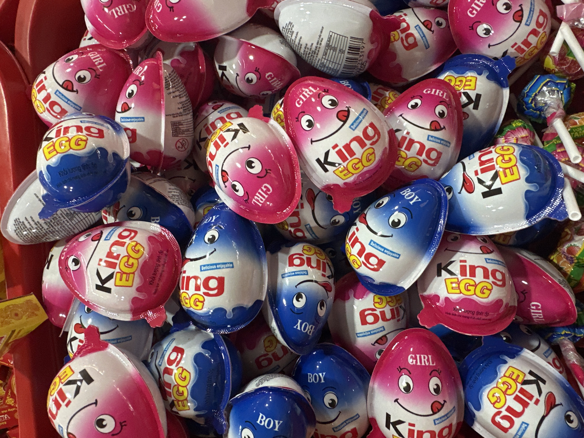 The image shows a close-up shot of a pile of "King Egg" chocolate eggs in a red bin. The eggs are individually wrapped and come in two versions: a pink one labeled "Girl" and a blue one labeled "Boy". Each egg has a cartoon face with a tongue sticking out. Text on the packaging is written in Vietnamese, indicating the products are being sold in Vietnam. The eggs are piled haphazardly, and some lollipops in wrappers are visible among them.