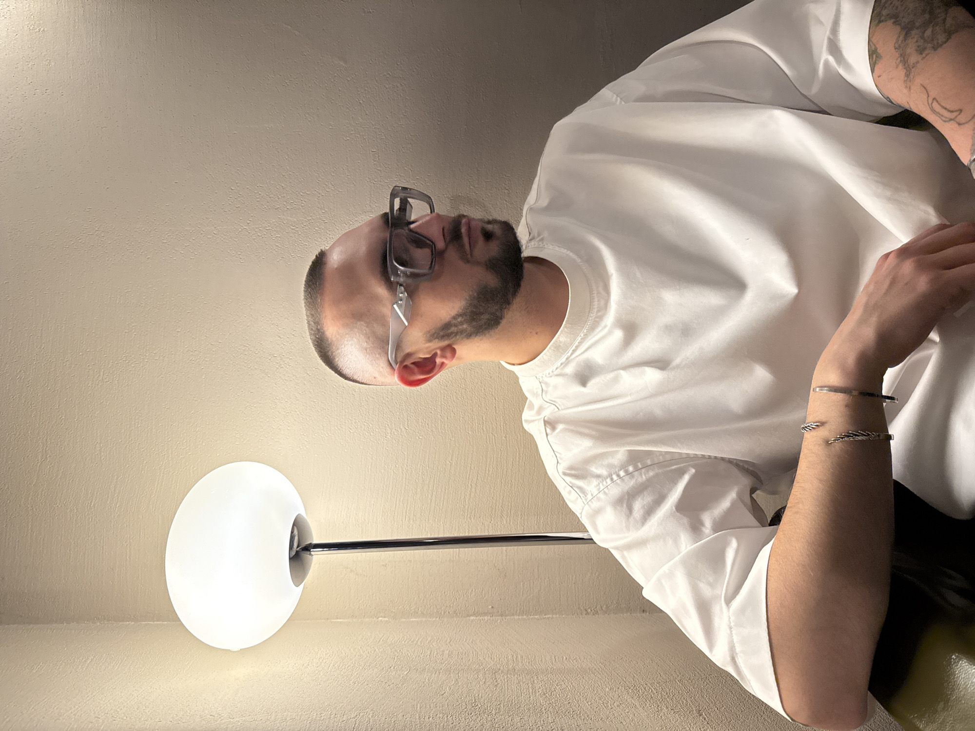 A medium, overhead shot shows a fair-skinned man with a trimmed beard and eyeglasses, wearing a white t-shirt and bracelets, seated next to a floor lamp with a glowing white globe. The background appears to be a neutral-toned wall. There is no indication of Yerevan, Armenia.
