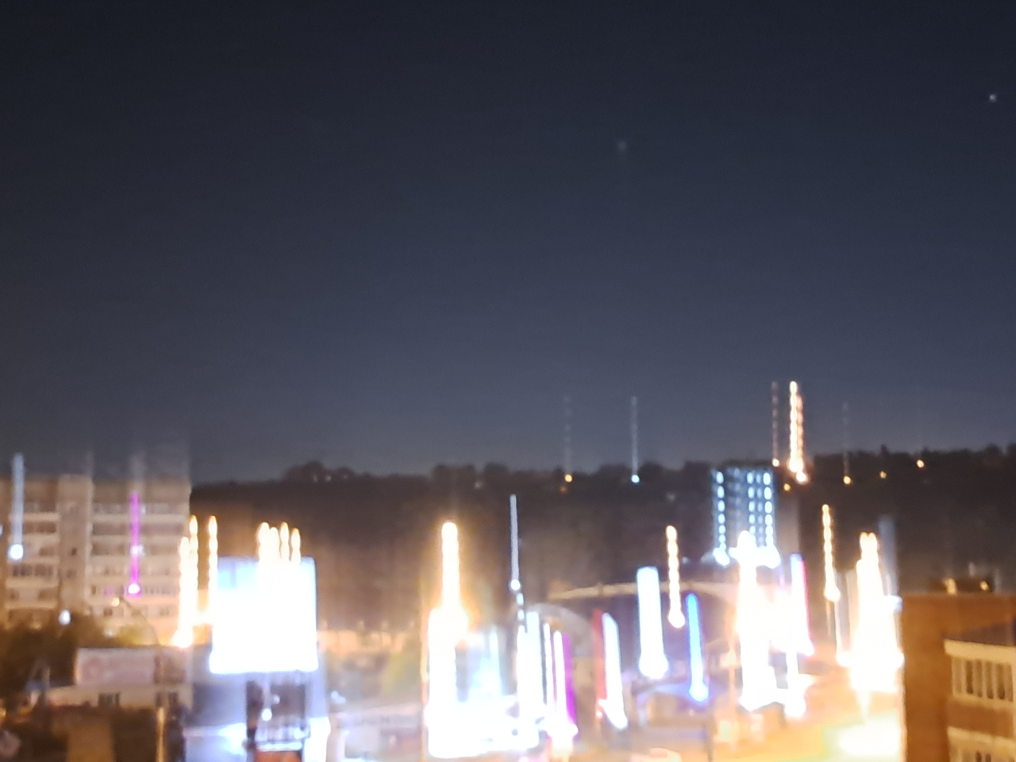 The image depicts a nighttime view of an urban landscape in Irkutsk, Russia. The scene is characterized by a dark sky with faint lights, possibly stars or distant aircraft. Below, the city is illuminated by various lights from buildings and decorative lighting installations. In the foreground, there are several buildings of varying heights, some with illuminated windows. A prominent feature is a series of tall, vertically oriented lights that appear to be part of a festive or decorative display, with some exhibiting pink and white illumination. In the middle ground, more illuminated buildings and structures are visible, including what appears to be a large, bright billboard or illuminated advertisement. The background consists of a dark, hilly terrain with several tall, slender structures that resemble communication towers, some of which are also lit. The overall impression is that of a city at night, possibly during a holiday or special event, with a focus on illuminated structures and a sense of urban activity despite the darkness.