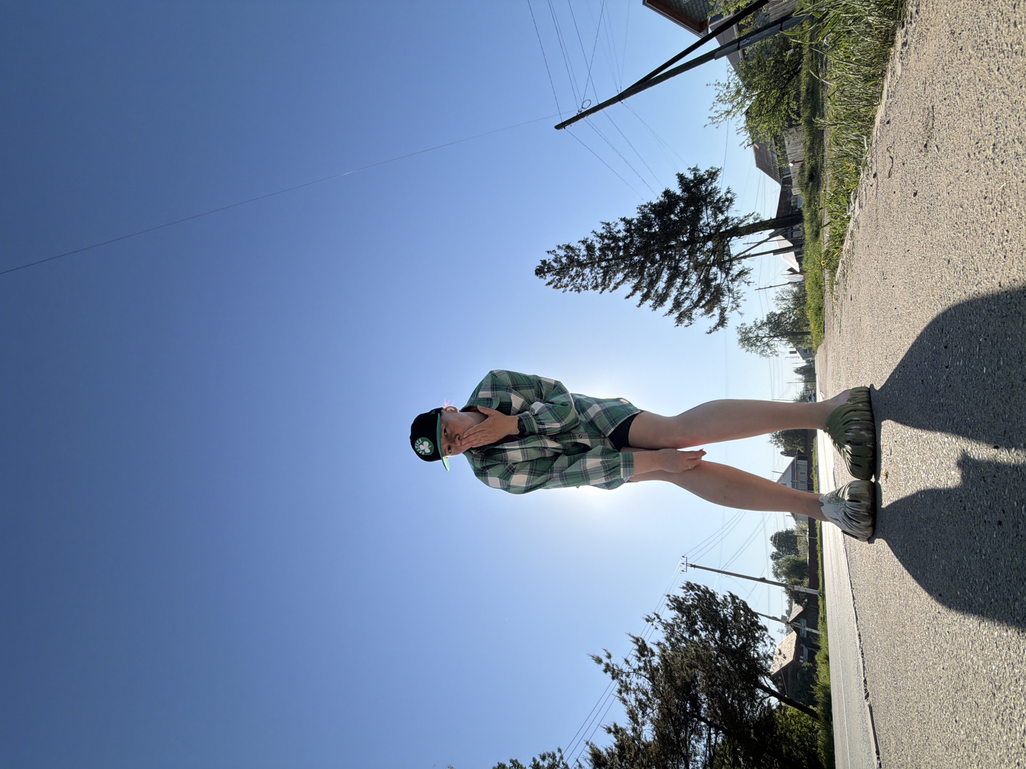 A young person wearing a green and white plaid shirt, black shorts, and green sandals with a Boston Celtics baseball cap is standing on a paved road in Chaikovsky, Russia. The person is holding their hands up to their face as if to shield themselves from the sun. The sun is behind them, causing a silhouette effect. Power lines run along the road. In the background, there are trees, grass, and houses. The sky is blue.