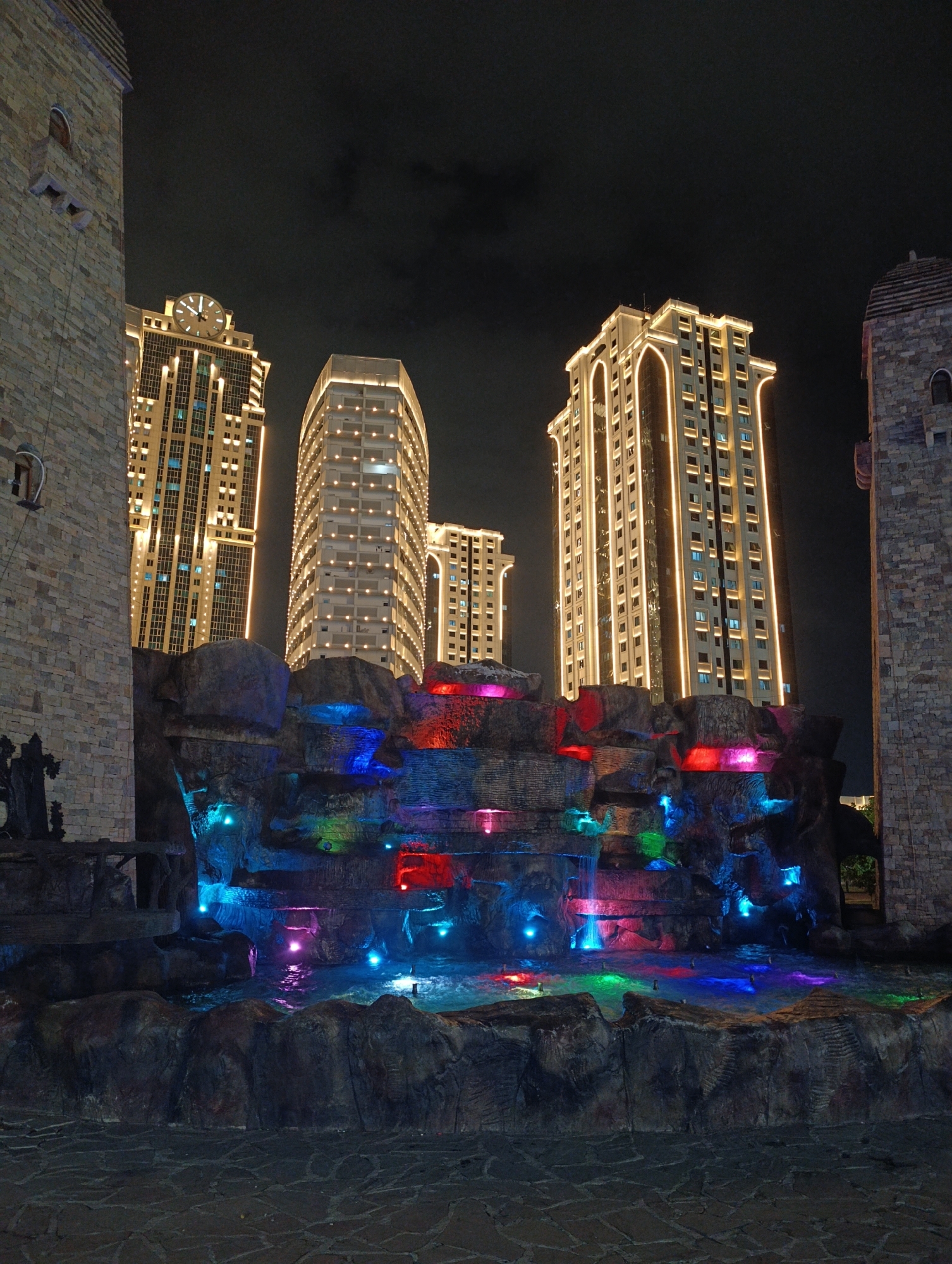 In the city of Grozny, Russia, at night, a man-made rock formation with a multi-colored illuminated waterfall is situated in front of several tall, modern buildings. The buildings are lit up with rows of lights, and the one on the left has a large clock face at its peak. The waterfall's water is illuminated with vibrant blues, greens, and reds, creating a dynamic visual display. The foreground consists of a paved area and two castle-like structures on either side of the waterfall, constructed from stone blocks. The sky above is dark and cloudy.