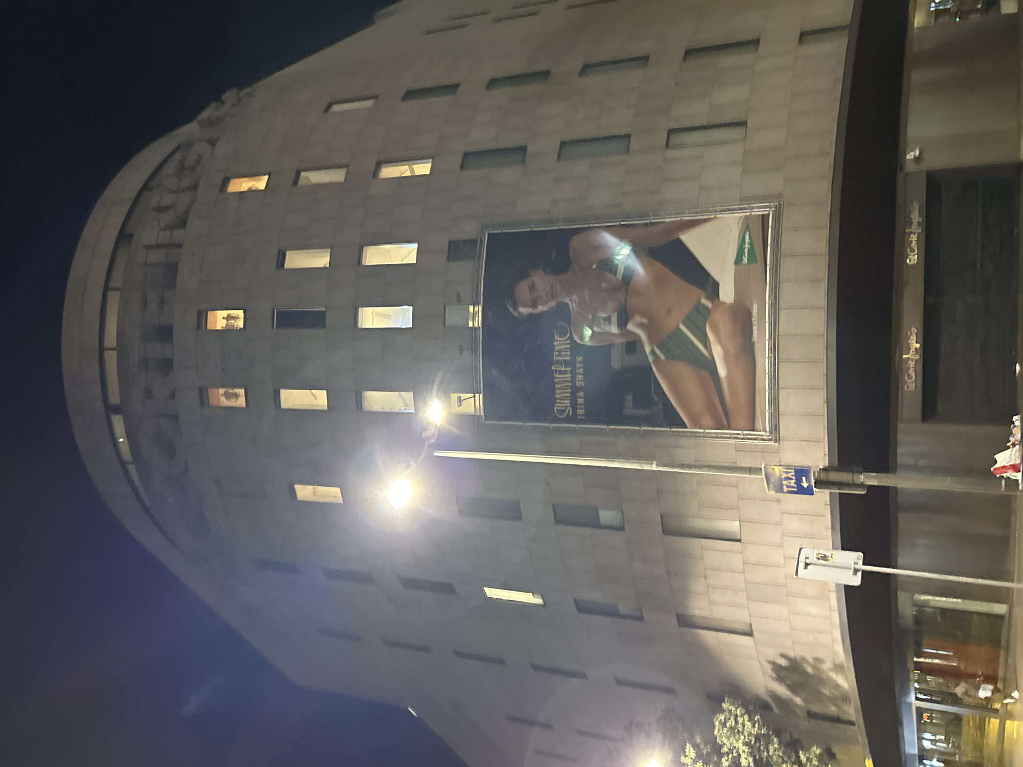 The image shows a building at night, likely located in Barcelona, Spain, based on architectural styles and signage. The building is tall and has a curved, cylindrical section on one side. Numerous horizontal rectangular windows are visible across its facade. A large advertisement featuring a woman in swimwear covers a significant portion of the building. Adjacent to the building's edge is a sign indicating a taxi stand. The overall lighting suggests a nighttime urban setting.