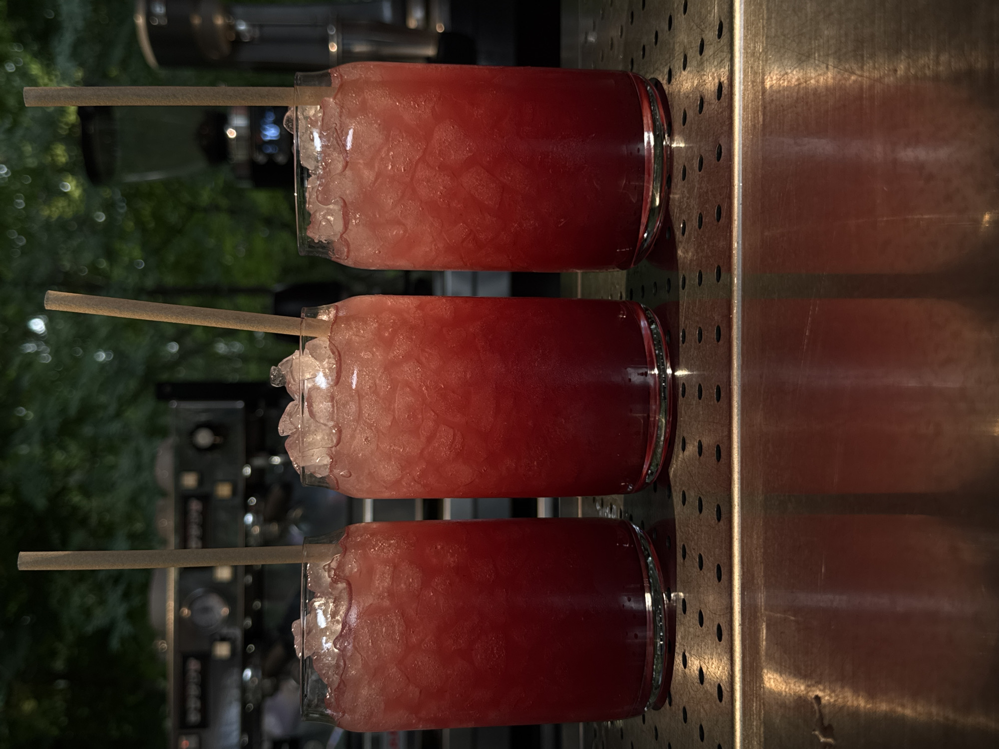 The image displays three tall glasses, each filled with a vibrant red or pink iced beverage and crushed ice, standing on a metallic bar counter. Each glass has a light brown straw, likely made from paper or bamboo, inserted into the drink. The beverages are arranged in a straight line, with their reflections clearly visible on the polished, solid section of the counter to their right. The counter also features a perforated metallic surface extending to the left of the glasses.
The background is softly blurred, revealing dark green foliage, suggesting an outdoor or semi-outdoor bar setting, possibly in Yerevan, Armenia. Behind the greenery, indistinct forms of bar equipment with subtle lights are visible, indicating an evening or nighttime scene. The overall lighting is dim, casting a focused glow on the refreshing beverages, which appear cool and inviting. There are no individuals present in the image, and no text is discernible.