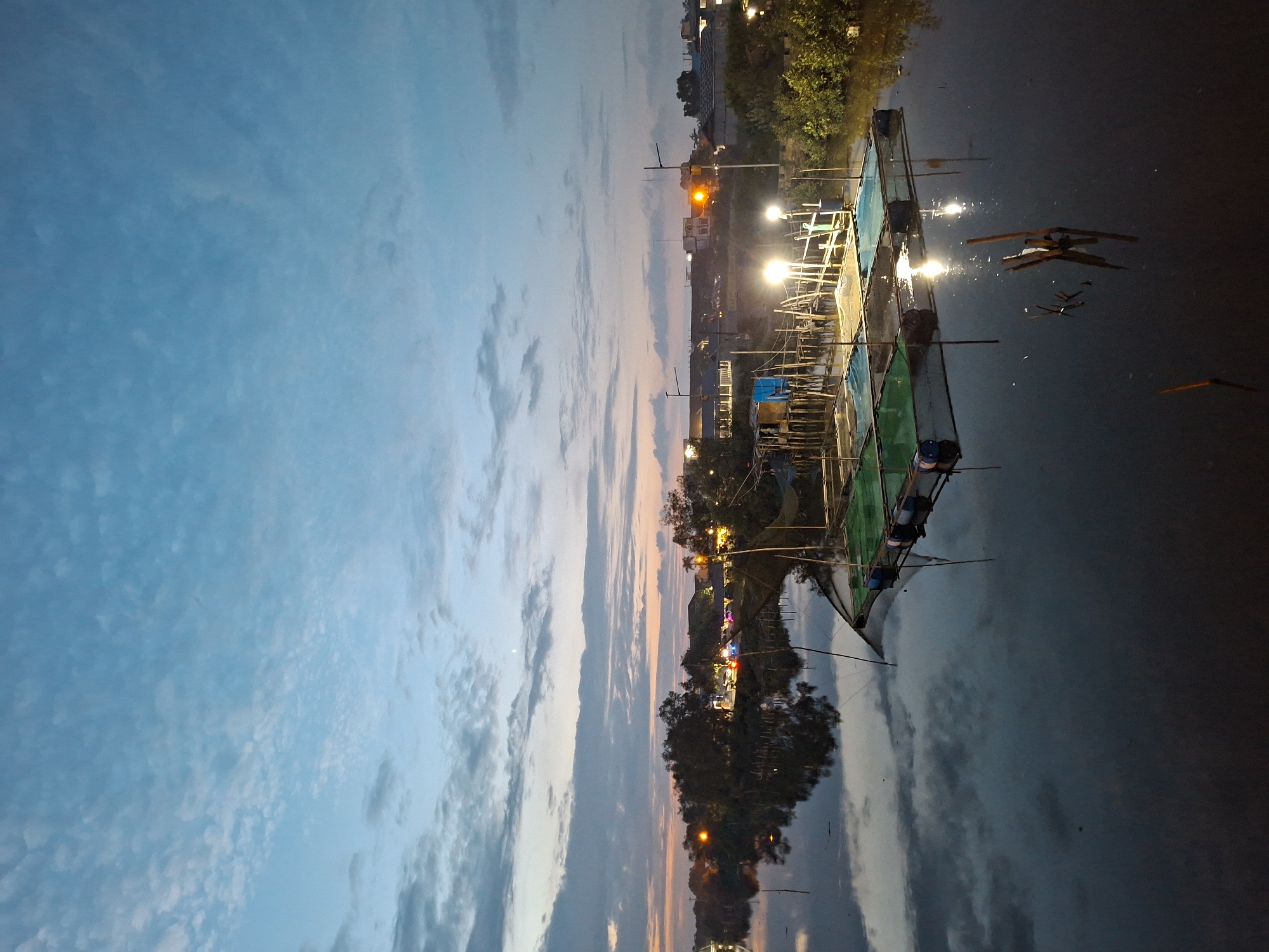 The image depicts a serene river scene at dusk or dawn in Lhokseumawe, Indonesia. The sky is a gradient of soft blue and pale orange, with wispy clouds scattered across it. The calm river water perfectly reflects the sky and the silhouettes of trees and distant buildings along the riverbank.
In the foreground, a floating structure, possibly a fish farm or a raft, is visible on the water. It is constructed with wooden poles and netting, with blue barrels used as flotation devices. Several bright lights illuminate parts of this structure, creating reflections on the water's surface.
Further back, along the riverbank, are trees, some with illuminated lights suggesting a settlement or businesses. The lights are a mix of yellow and white, casting a warm glow. The overall impression is one of tranquility and stillness, characteristic of a quiet evening or early morning by the water. There are no people visible in the image.