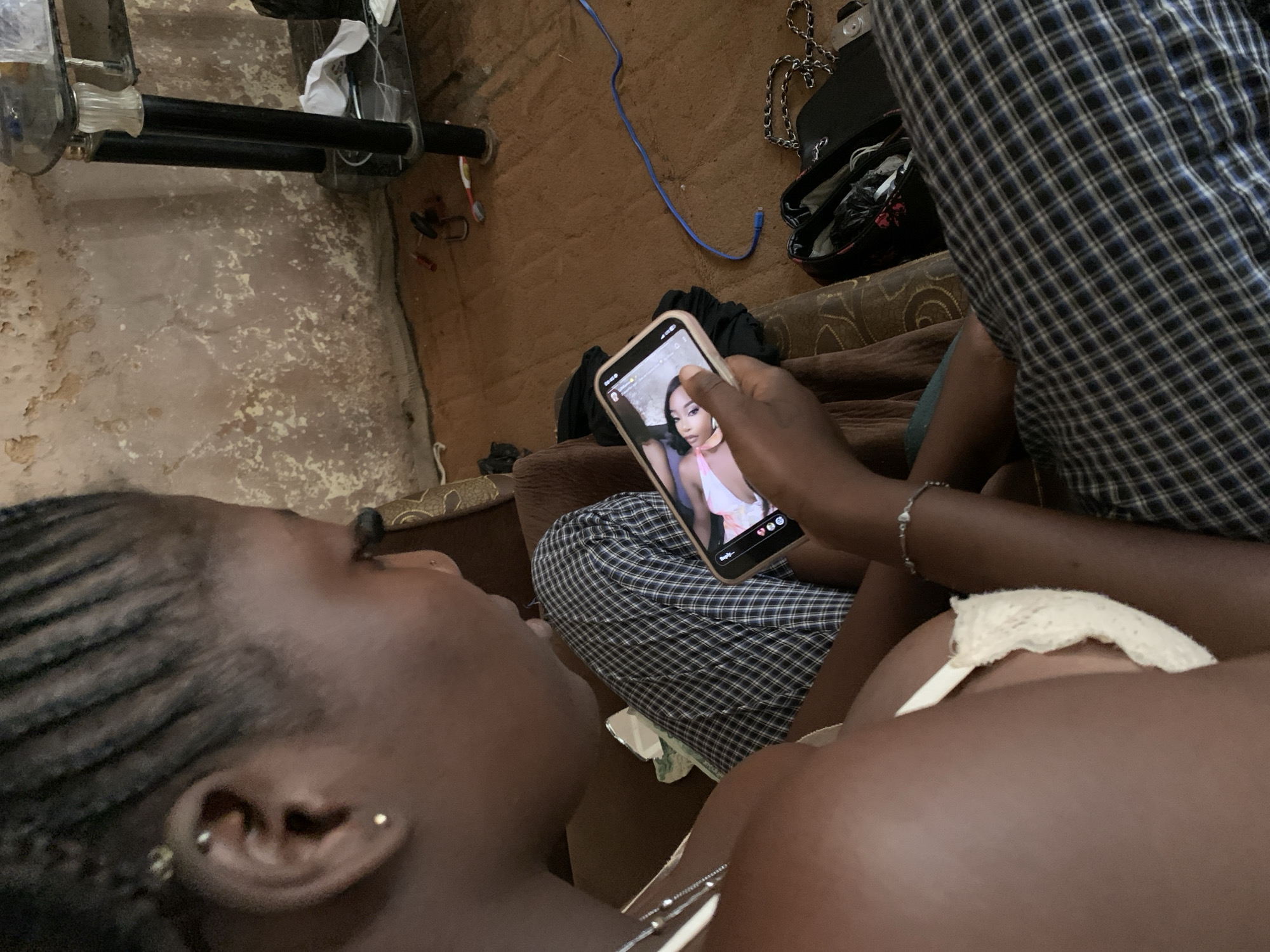 The image shows a close-up perspective of two individuals in what appears to be a dimly lit indoor setting, possibly a living room. The foreground is dominated by the head and shoulder of a person whose face is turned away from the camera, revealing braided hair and an earring. This person is holding a smartphone, displaying an image of another individual, a young woman with dark hair and a pink top.
The phone's screen shows a social media interface with engagement icons like hearts and speech bubbles. The hand holding the phone has a bracelet on the wrist. In the background, another person is partially visible, wearing a checkered shirt. The surroundings include a floor with a textured, possibly carpeted surface, and some discarded items, including a blue cable. There are hints of furniture and possibly a bag.
The overall mood seems relaxed, with the focus on the shared viewing of the phone screen. The lighting suggests it could be evening or nighttime, with no indication of the weather. The mention of Jos, Nigeria provides a geographical context for the scene. The image is factual in its depiction of the subjects and their immediate environment.