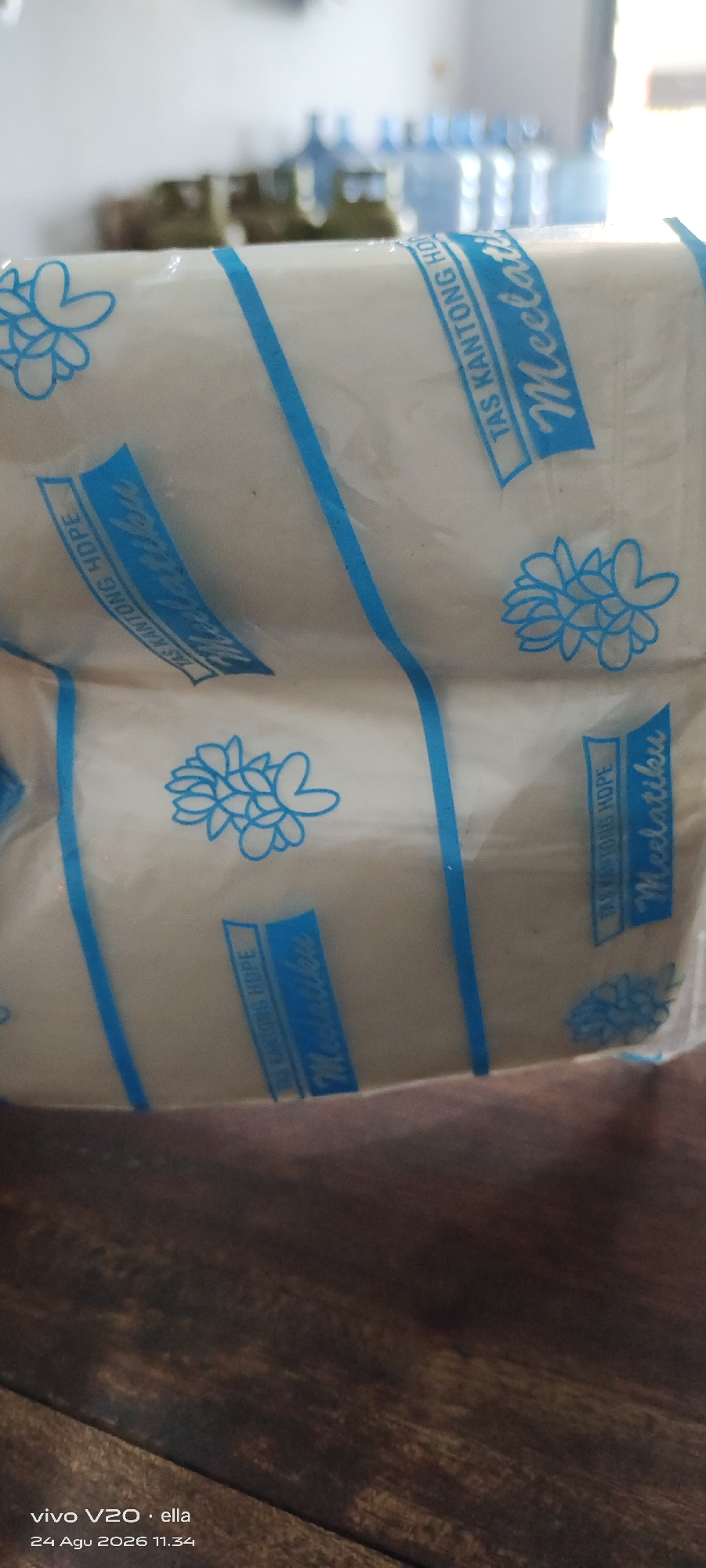The image displays two rolls of white plastic bags, each wrapped in transparent plastic. A blue band encircles each roll, featuring a stylized flower design and text. The text on the bands reads "TAS KANTUNG HDPE" followed by the word "MelatiKu" in a script font. The rolls are placed on a dark wooden surface. In the background, out of focus, are several blue plastic water bottles. The bottom left corner of the image shows text indicating a mobile device model "vivo V20" and a date "24 Agu 2026 11.34". The scene is likely an indoor environment, possibly a shop or storage area, in Simo, Indonesia.