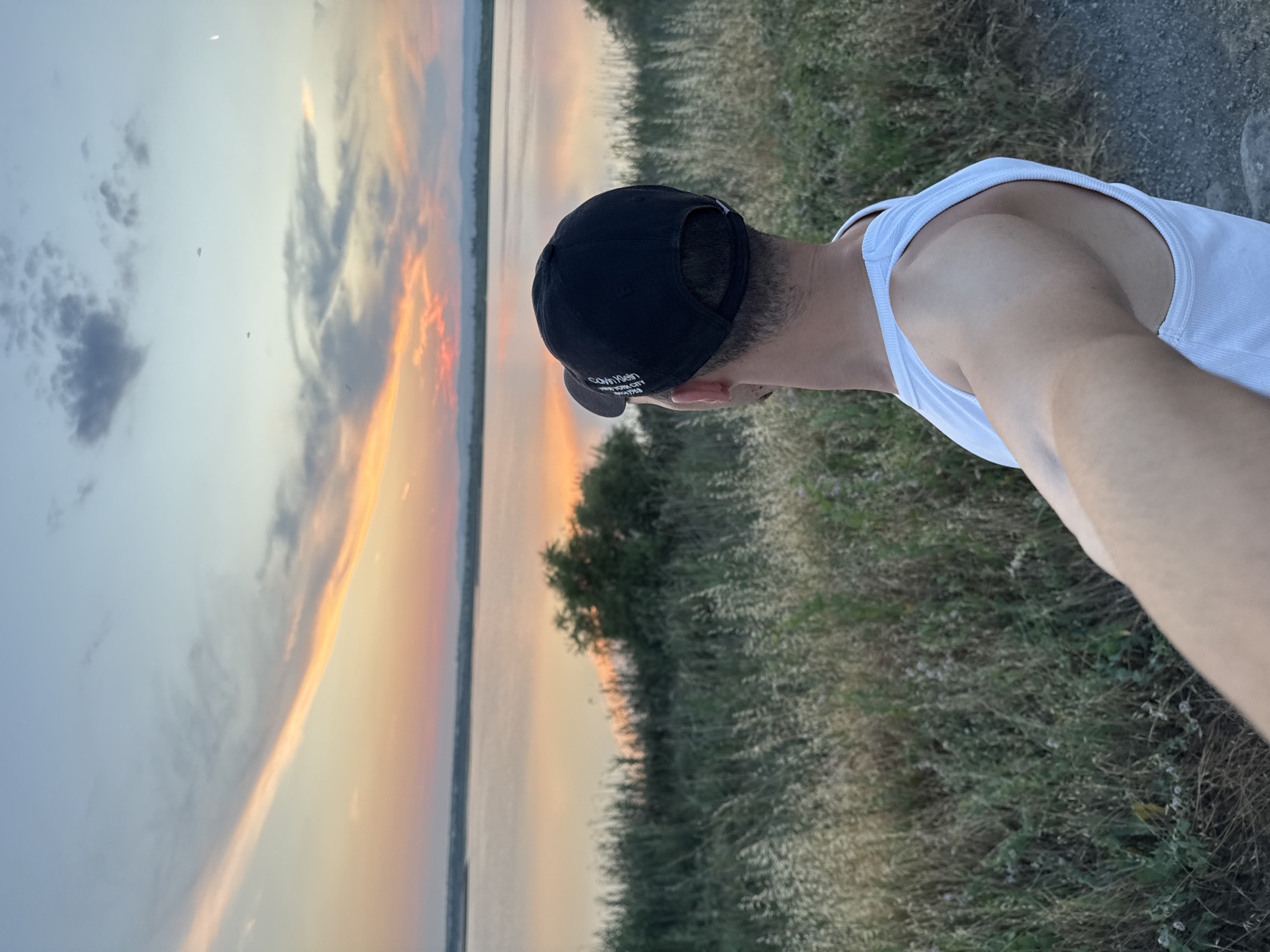 In the city of Pomorie, Bulgaria, a man wearing a white tank top and a black baseball cap stands facing a large body of water at sunset. The sky is filled with hues of orange, pink, and blue, with scattered clouds. The water reflects the colorful sky, creating a serene scene. The man is holding his arm out, presumably to take a selfie. The foreground is composed of dry grass and vegetation, suggesting a natural, undeveloped area. The background shows a distant shoreline with hills. The overall atmosphere is peaceful and contemplative.
