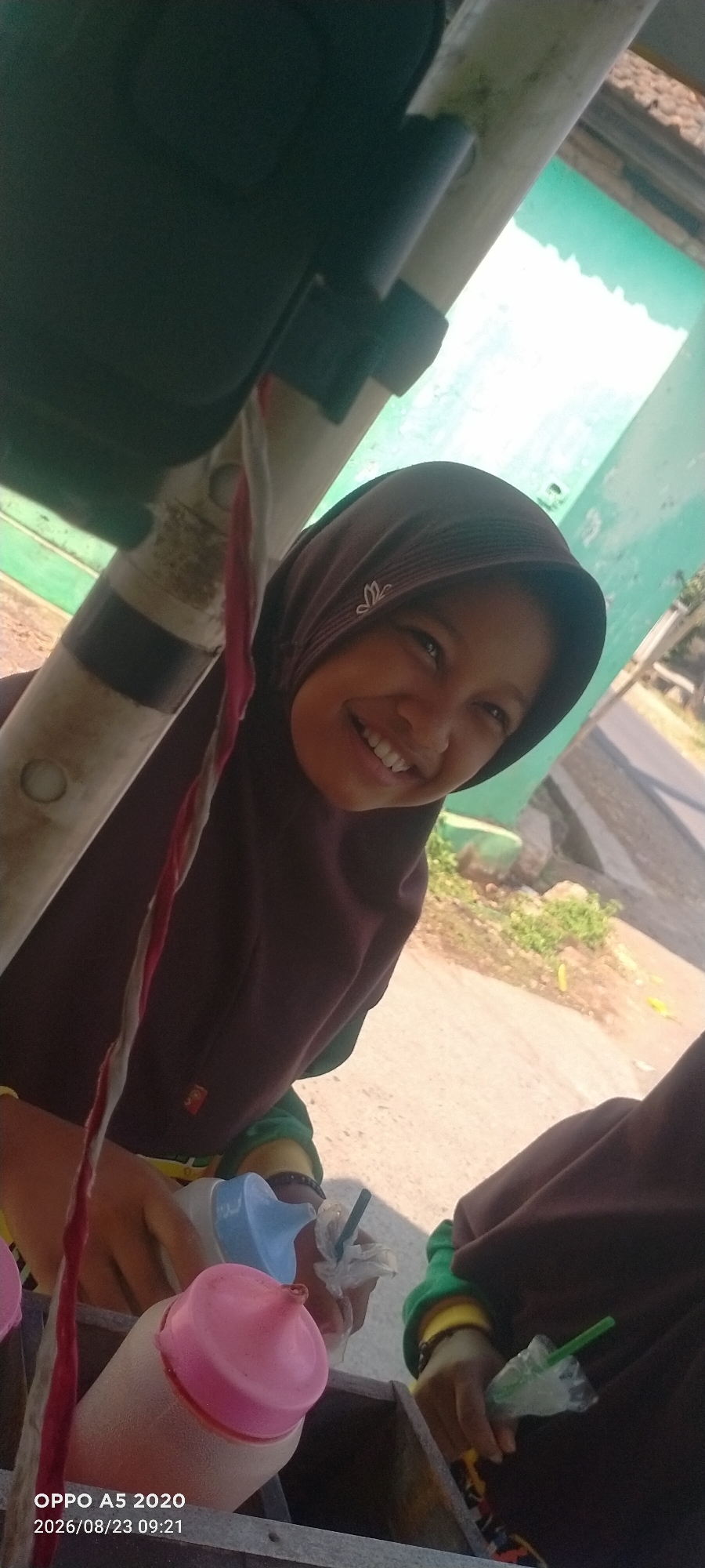 A young girl wearing a brown hijab smiles as she poses for a selfie. She is sitting in a wooden cart, holding a pink-capped bottle in her left hand and a blue-capped bottle with a straw in her right. The cart appears to be on the side of a road with some vegetation and a building in the background. The timestamp on the image indicates it was taken on August 23, 2026, at 09:21 in Sowan Kidul, Indonesia.