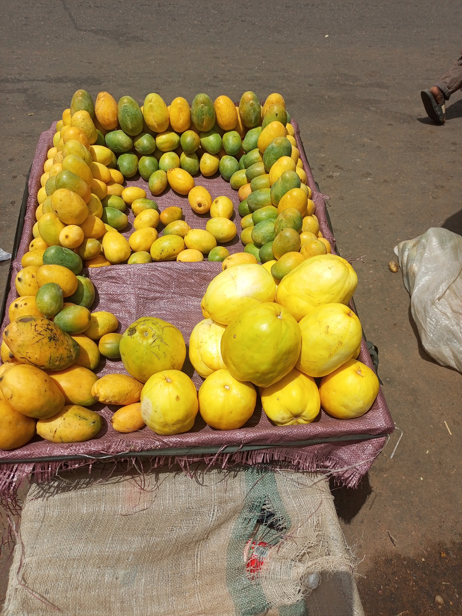 A street vendor's cart in Tempure, Nigeria, displays a variety of mangoes and yellow citrus fruits for sale. The cart is covered with a pink mat, on top of which are arranged rows of fruits, varying in color from green to yellow. The fruits are arranged in a structured manner. In the background is a paved street. A leg of a person wearing sandals is visible on the right. A bag of white material is also located on the right. The cart is supported by a beige bag.