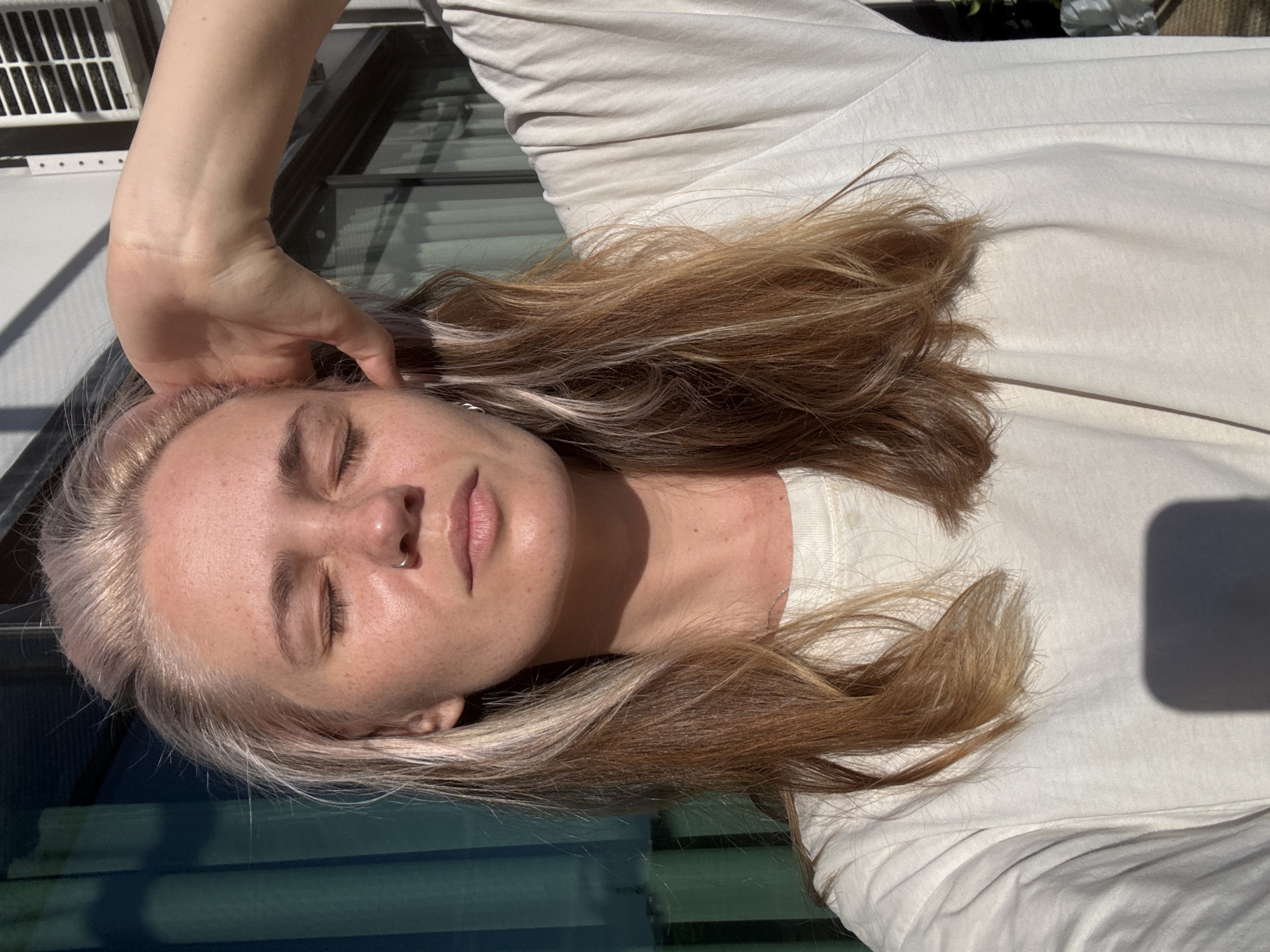In a medium shot, a young Caucasian woman with blonde hair is positioned against a backdrop of large glass windows and a white air conditioning unit. Her eyes are closed, and her right hand is raised, touching the side of her head. She is wearing a loose-fitting white t-shirt, and a shadow is cast on her chest. The lighting suggests an outdoor setting, possibly a balcony or patio. There is no indication of the specific location within Choeng Thale, Thailand.